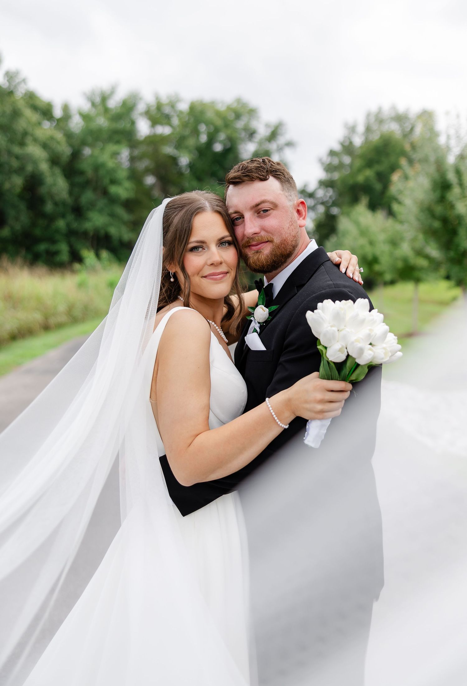 Bride and groom embrace outdoors, her white veil flowing, holding white tulips, with lush green trees in the background.At The Venue at Birc