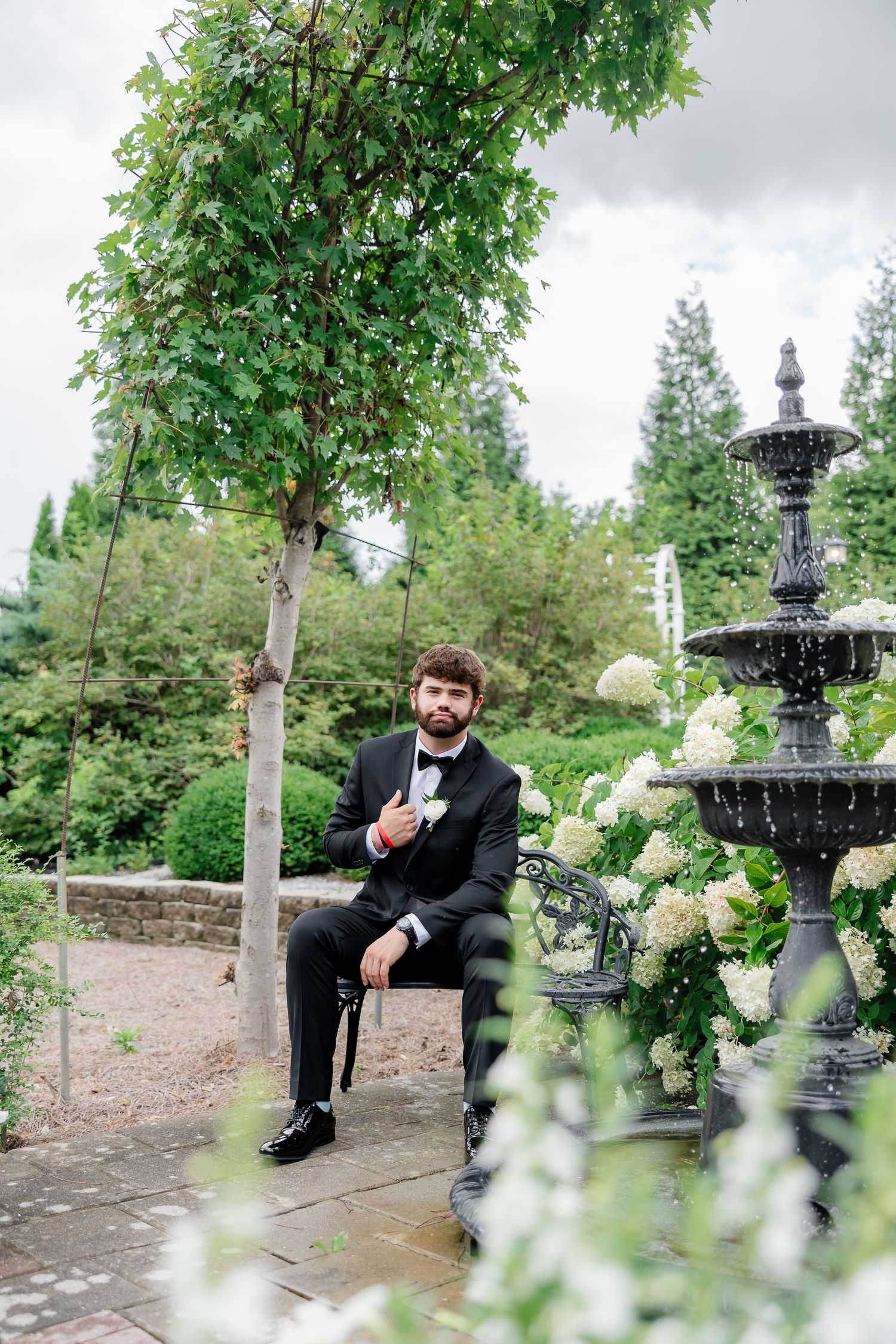 Groom in black tuxedo sitting on bench near fountain and white hydrangeas in lush garden setting.