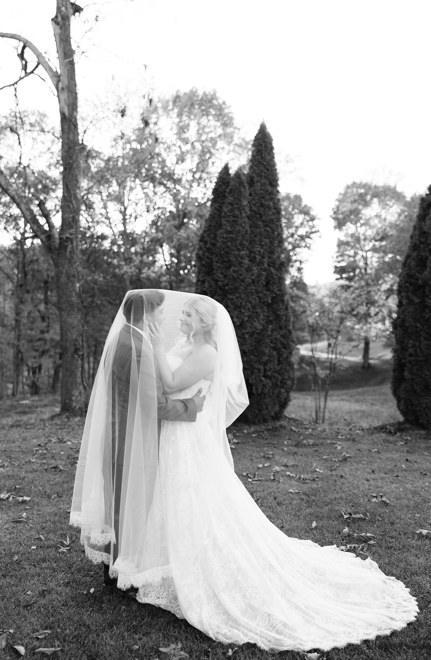 Black and white photo of a bride in a flowing gown and veil standing outdoors among tall cypress trees.