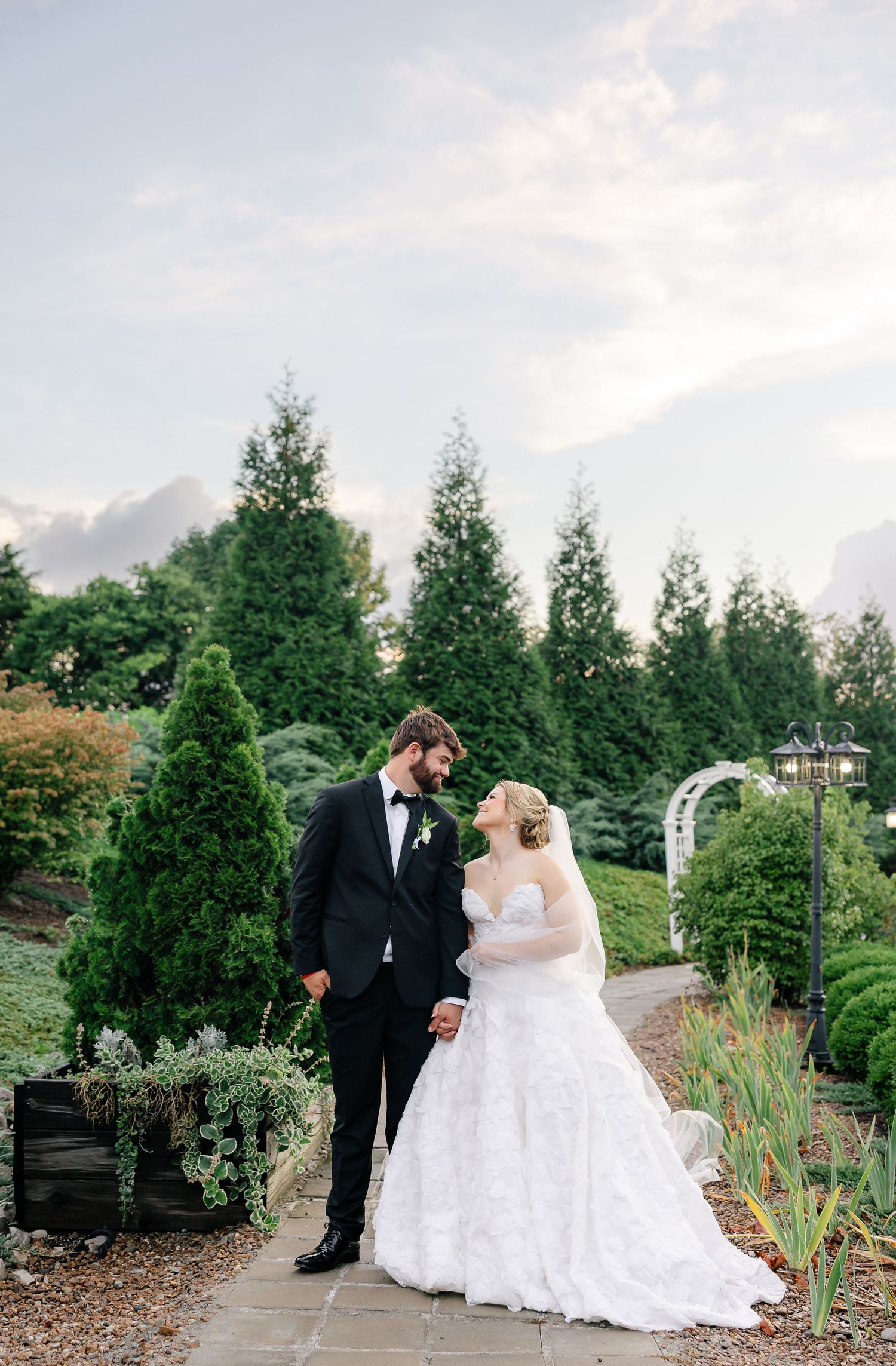 Bride and groom share a romantic moment in a lush garden setting, with the bride in a lace gown and groom in a black tuxedo.