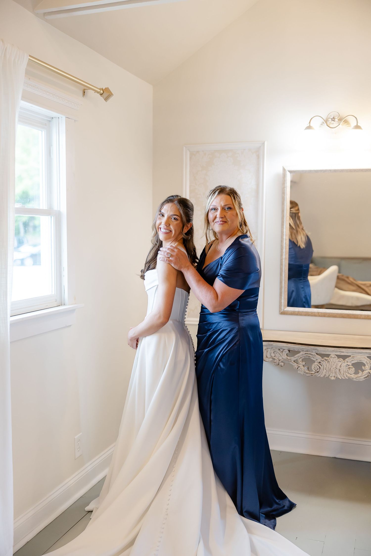 Bride in white gown with woman in navy blue dress posing together in elegant bridal suite near window.
