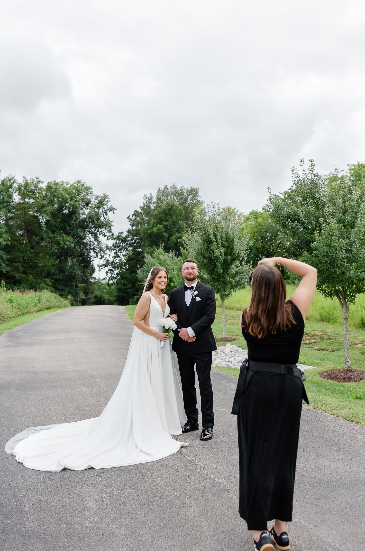 Photographer capturing bride in white gown and groom in black tuxedo posing on a tree-lined path.