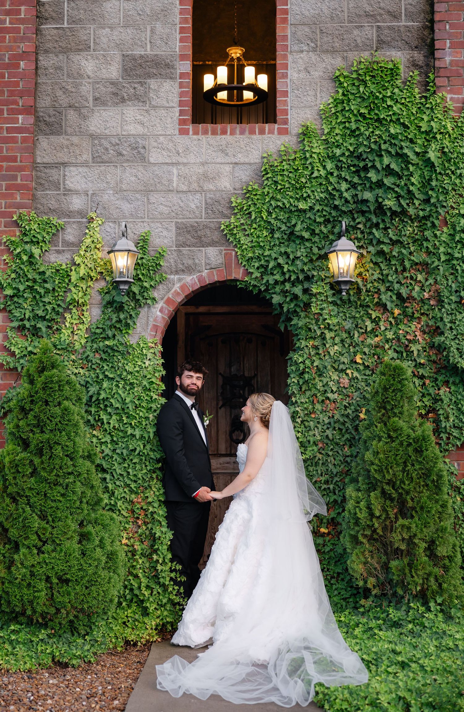 Bride and groom hold hands before ivy-covered stone church entrance with lanterns on their wedding day.