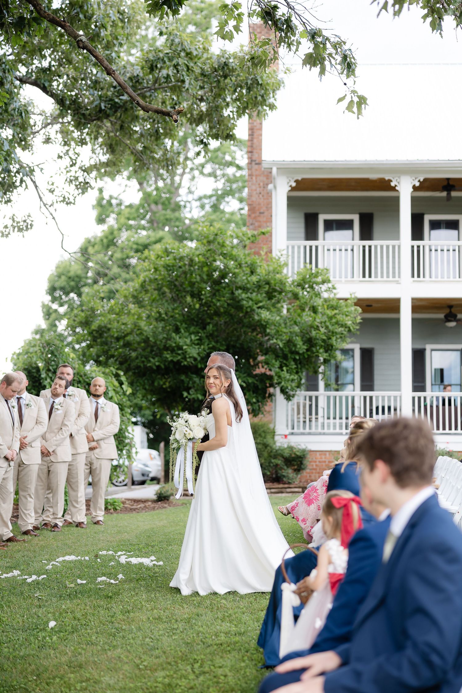 Bride walks down outdoor aisle in white gown, groomsmen in tan suits, historic house backdrop.
