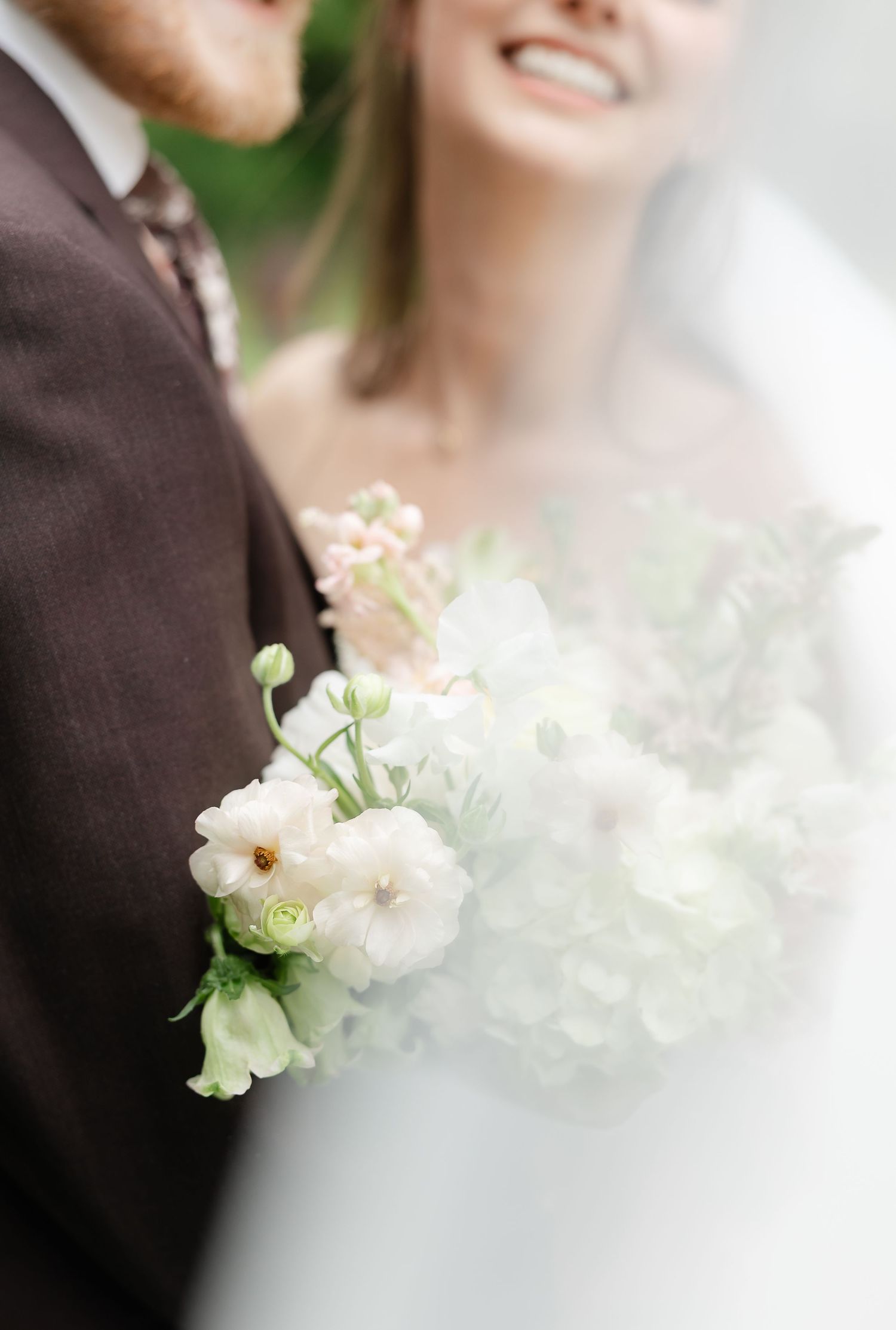 Bride holding a soft white and blush wedding bouquet with ranunculus and hydrangeas beside groom in brown suit.