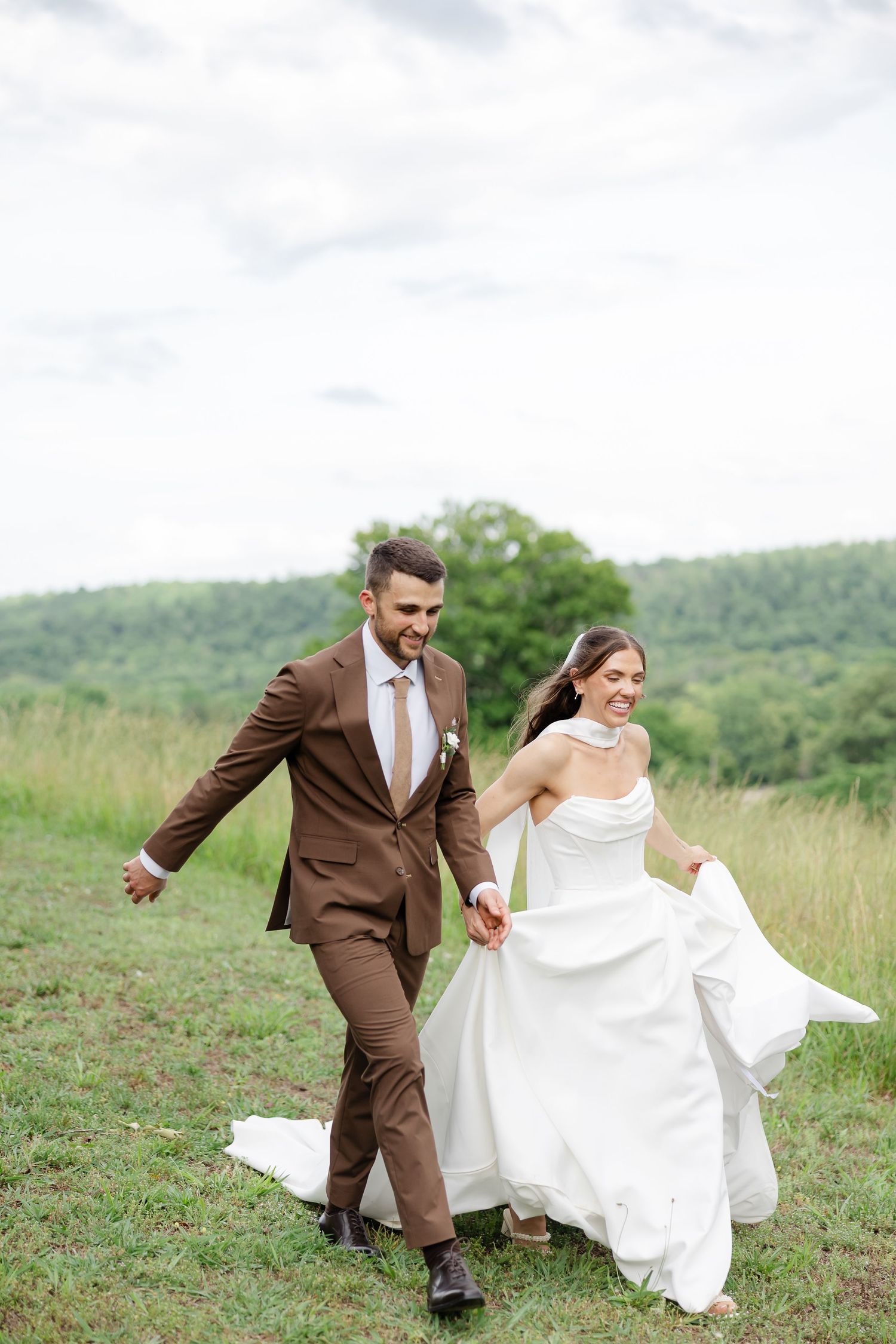 Smiling bride and groom holding hands running through green meadow, bride in white gown, groom in brown suit.