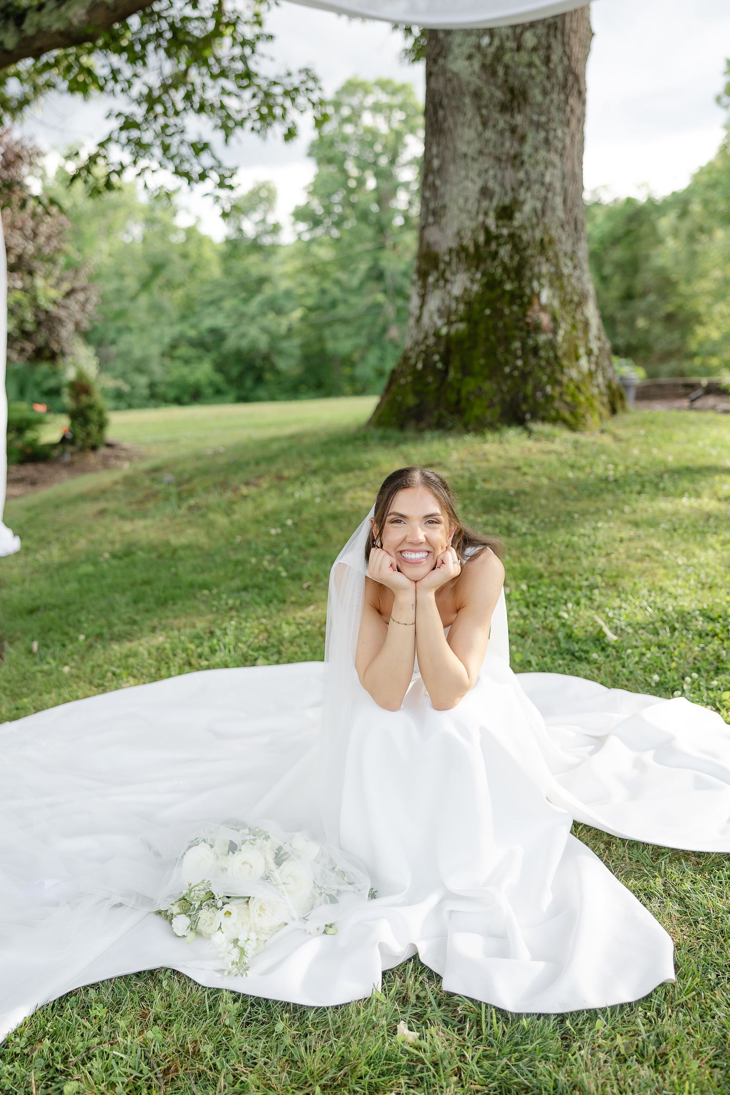Smiling bride in white gown and veil sits on grass with bouquet, posing with hands under chin near large tree.