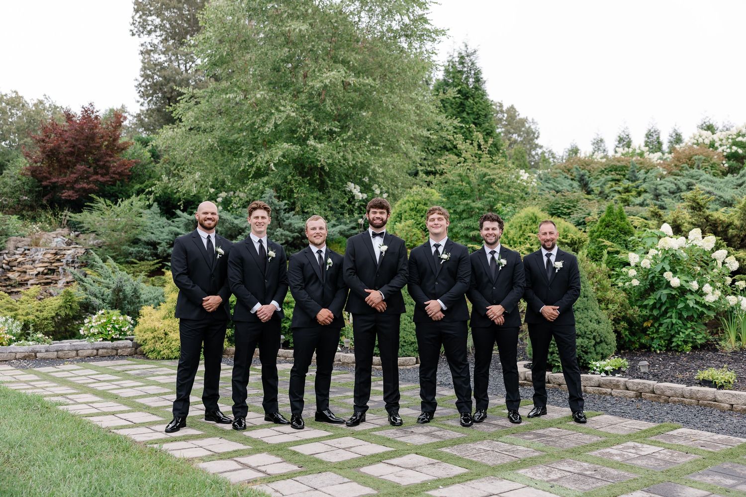 Seven groomsmen in matching black suits pose together on a garden pathway surrounded by lush greenery.