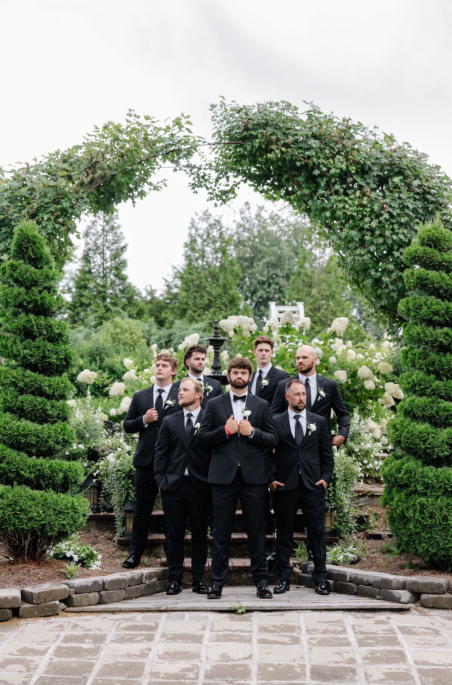 Groom and groomsmen in black tuxedos pose under a lush green garden arch with white floral arrangements.