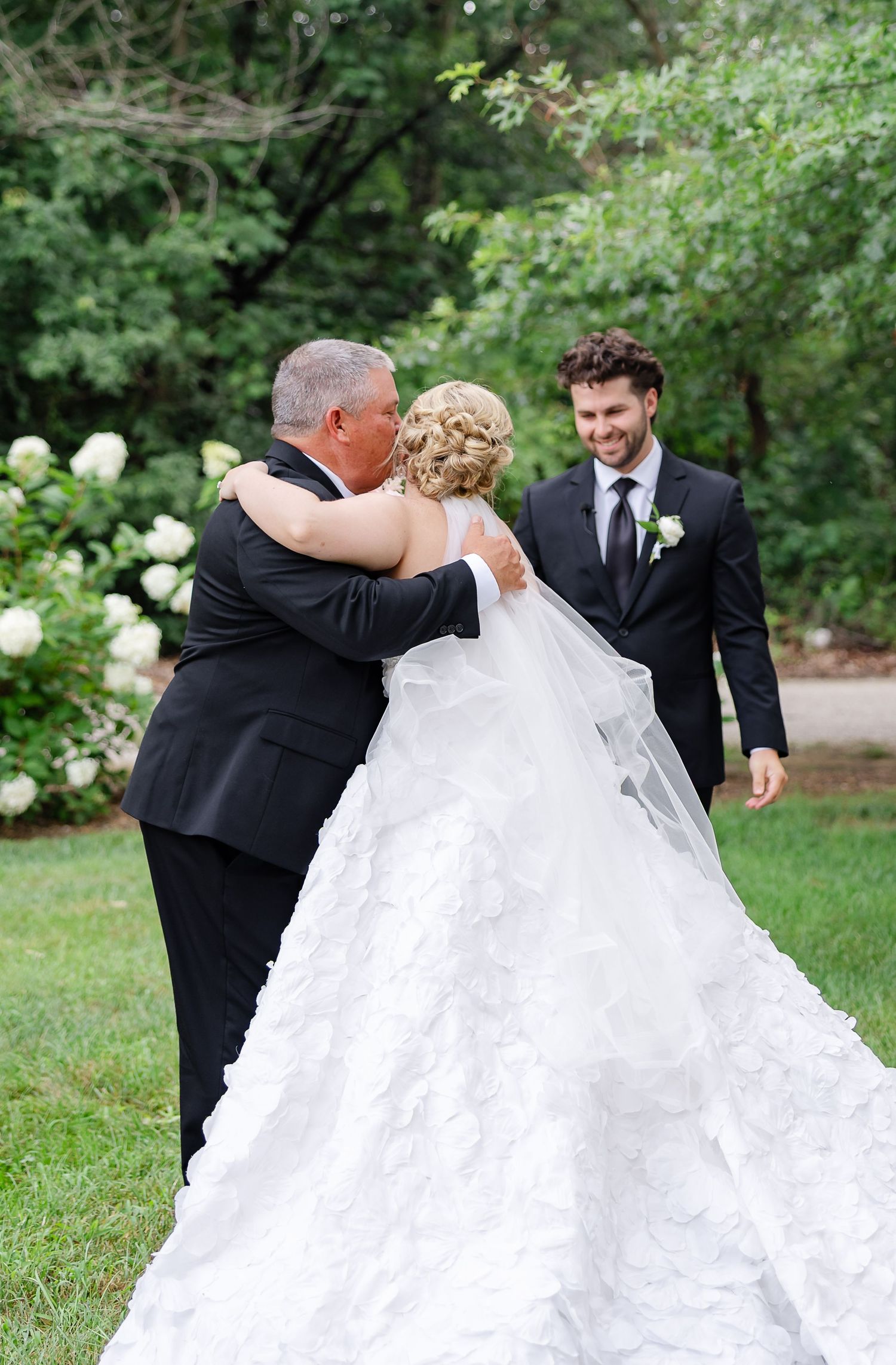 Father embracing bride in white ruffled gown as smiling groom watches during outdoor wedding ceremony.