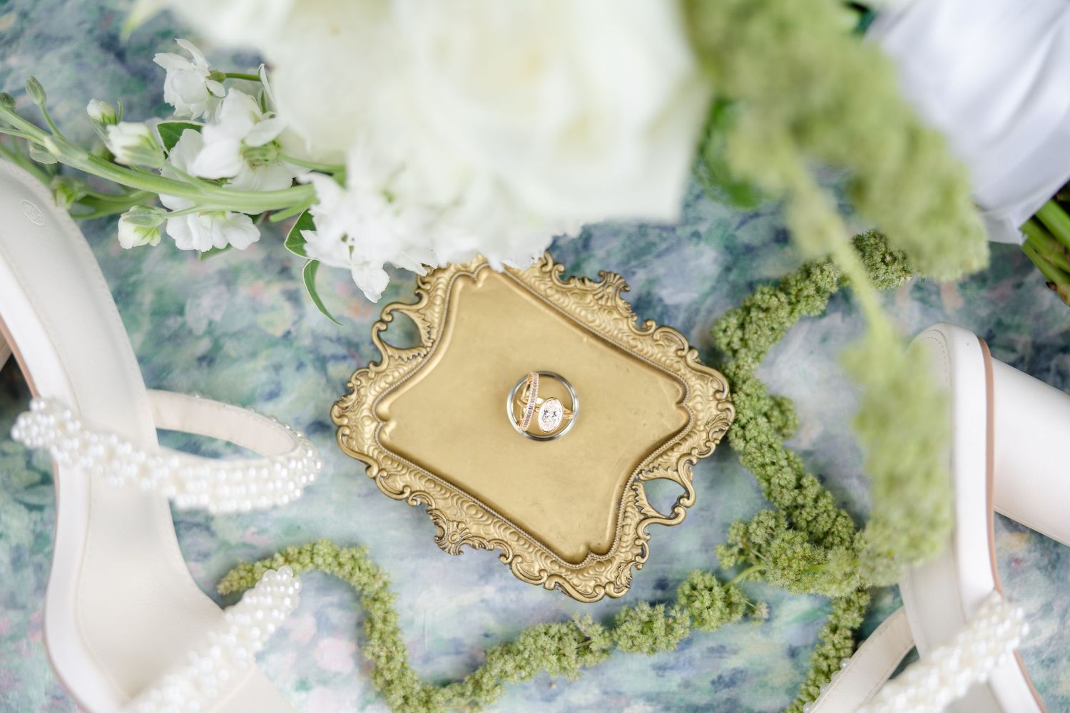 Gold ornate ring holder with diamond engagement ring on blue floral fabric with white flowers and pearls.