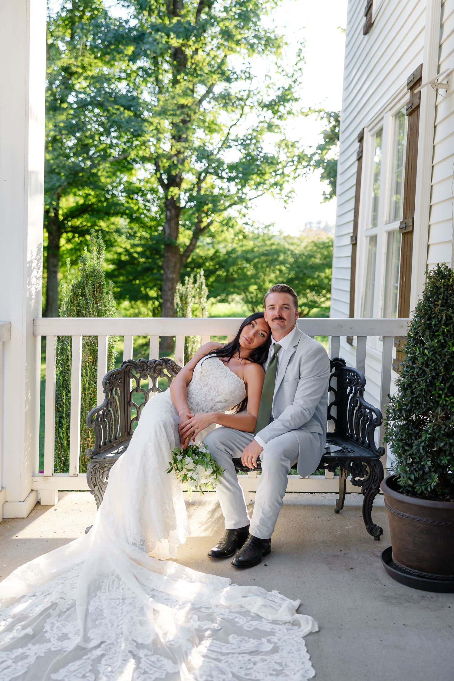 Bride and groom sitting together on a porch bench, smiling, surrounded by lush greenery at their wedding venue.At Drakewood Farms