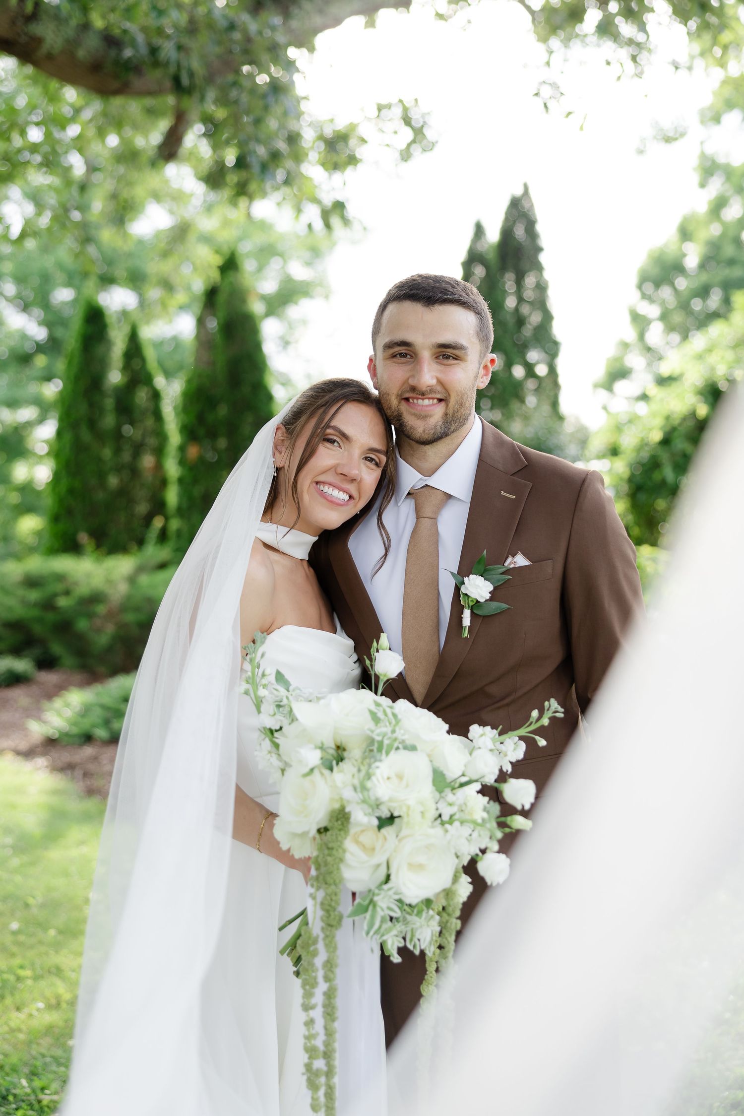 Smiling bride and groom pose outdoors, bride holding white floral bouquet, groom in brown suit with boutonniere.