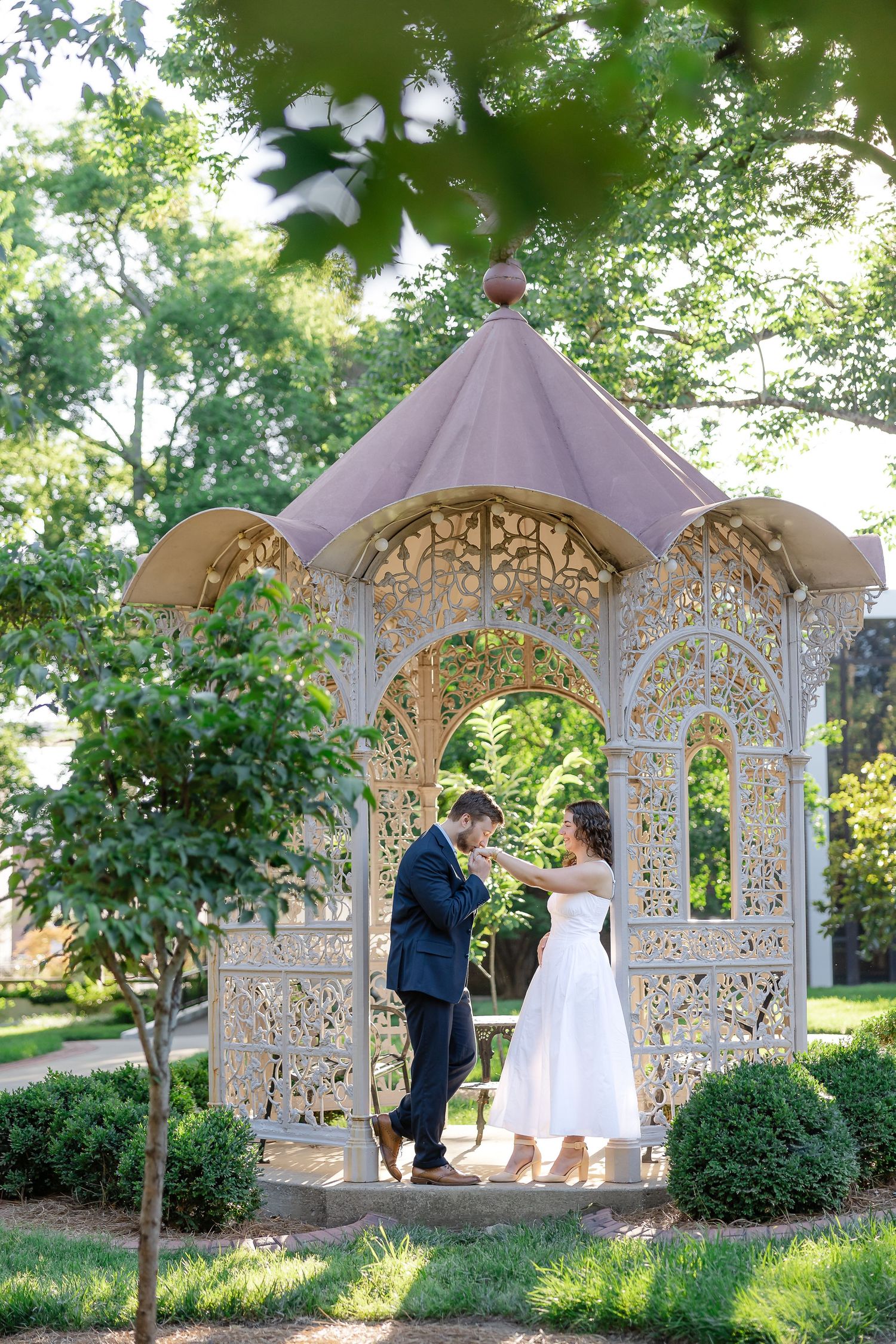 Couple sharing a romantic moment under an ornate white gazebo surrounded by lush green trees in a garden setting.