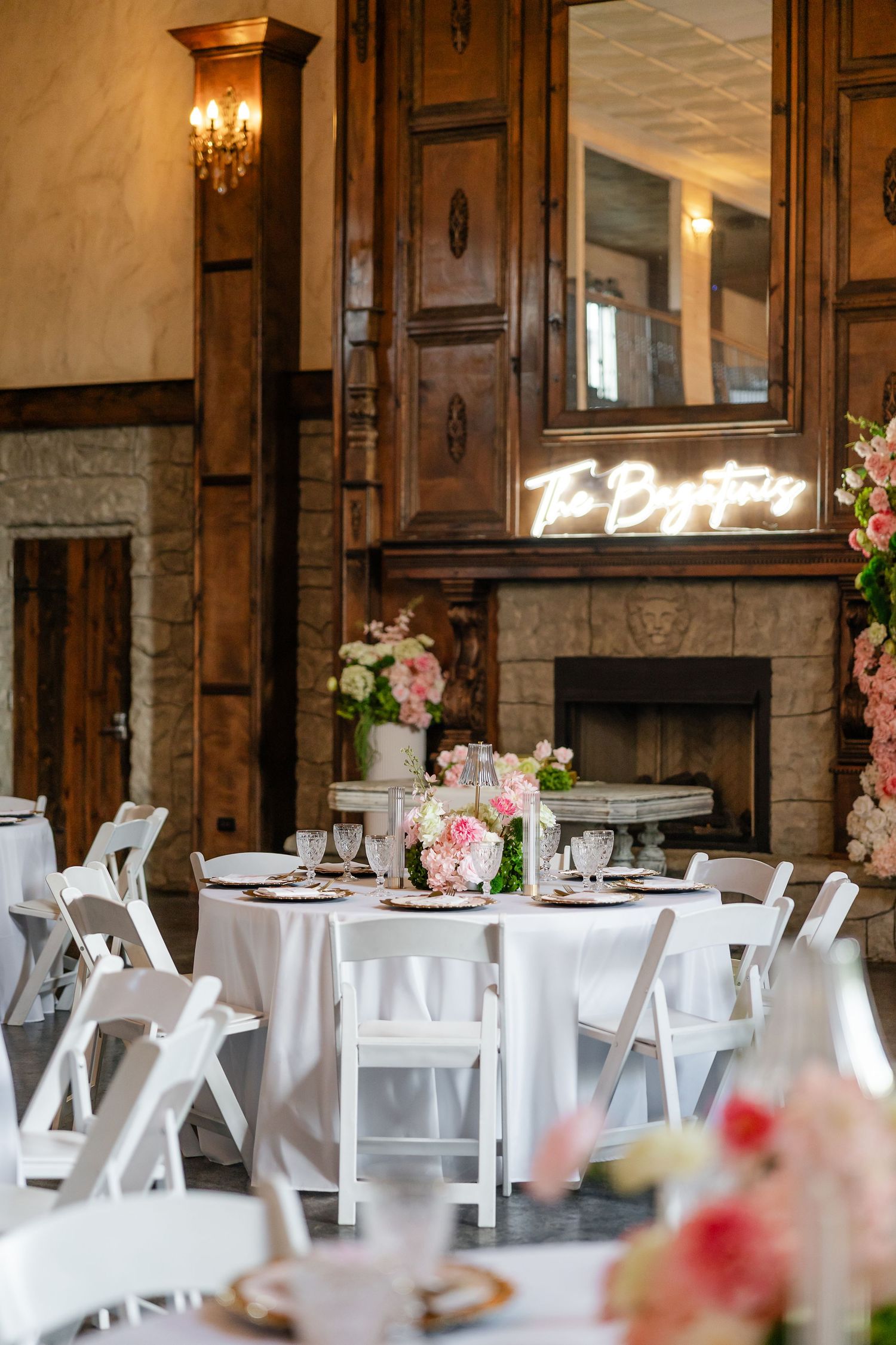 Elegant wedding reception with white chairs, floral centerpieces, and a neon sign above a stone fireplace.
