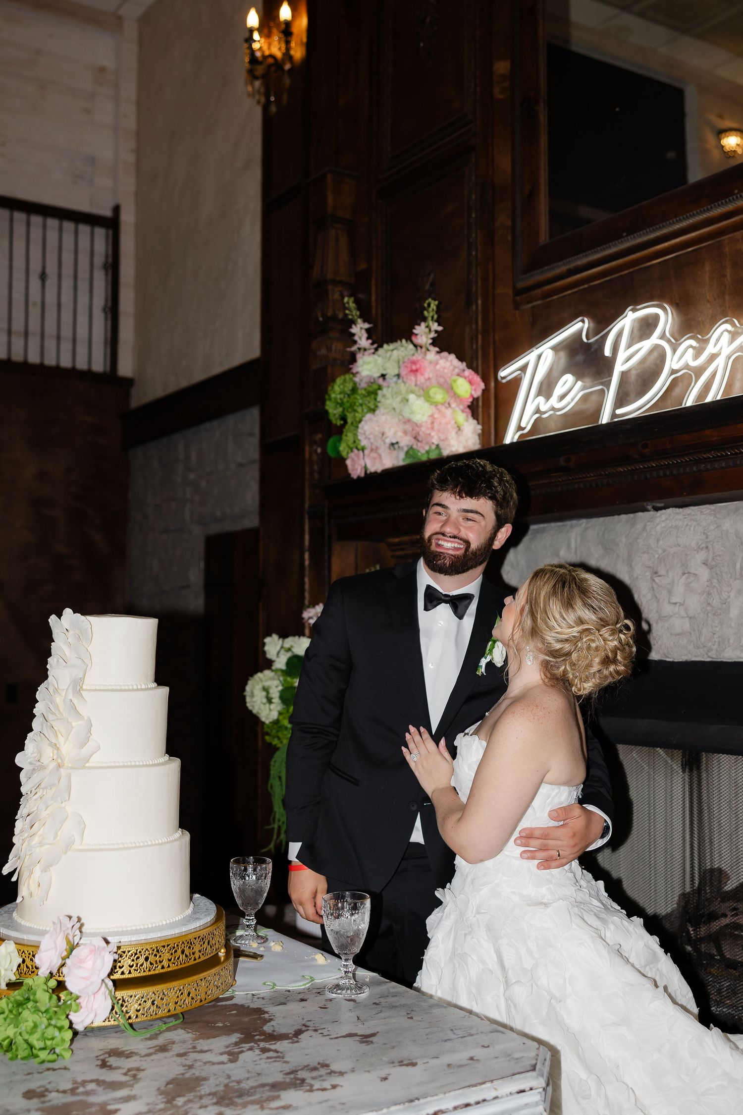 Bride and groom smile together beside a white tiered wedding cake at elegant reception venue with neon bar sign.