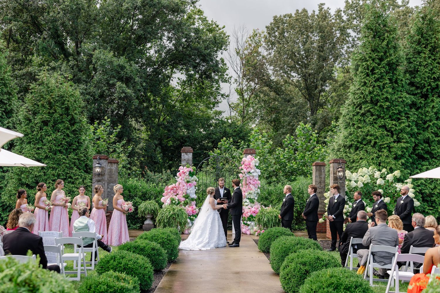 Outdoor garden wedding ceremony with bride and groom at floral arch, bridesmaids in pink, guests seated on white chairs.