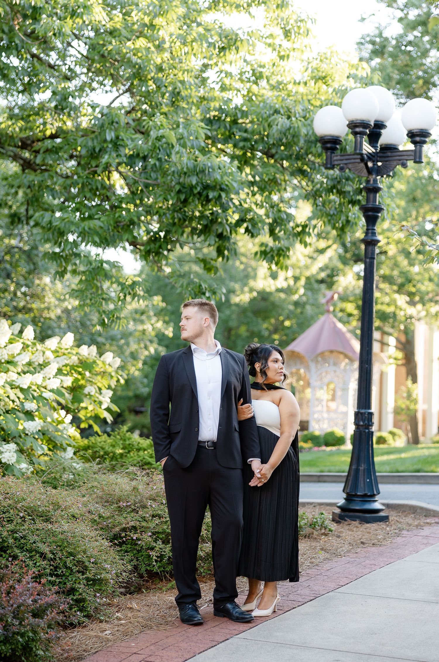 Couple in black and white formal attire holding hands by a decorative lamp post in a lush green park setting.