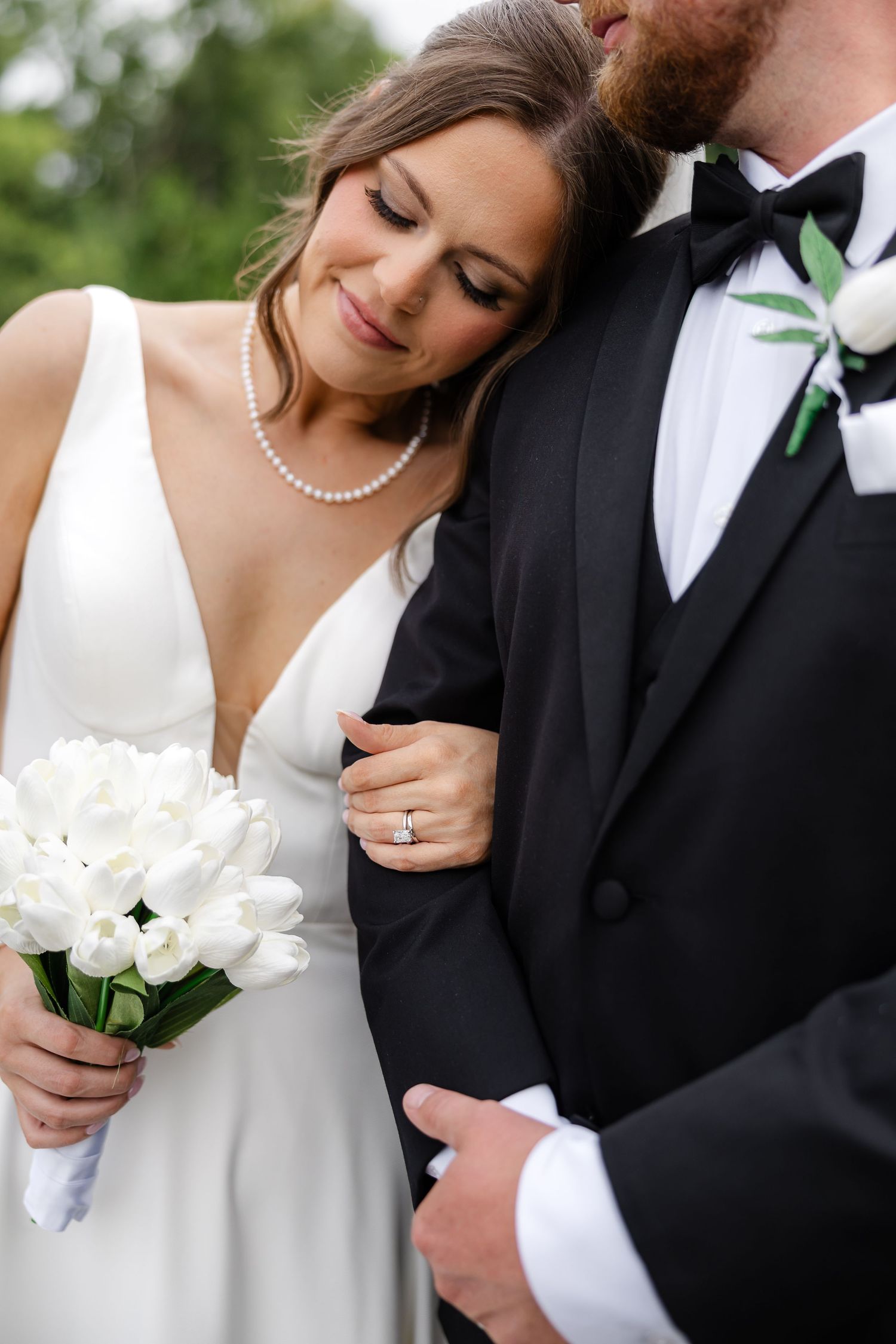 Bride in white gown holds tulip bouquet, leaning on groom in black tuxedo with bow tie and boutonniere.