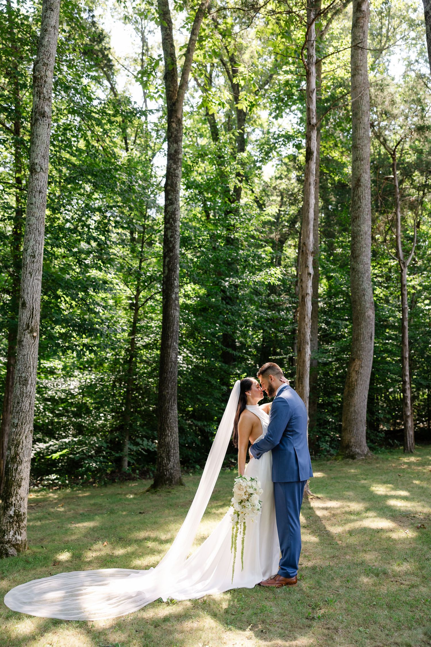 Bride and groom share a romantic kiss outdoors, surrounded by tall green trees, with a flowing cathedral veil trailing on the grass.
