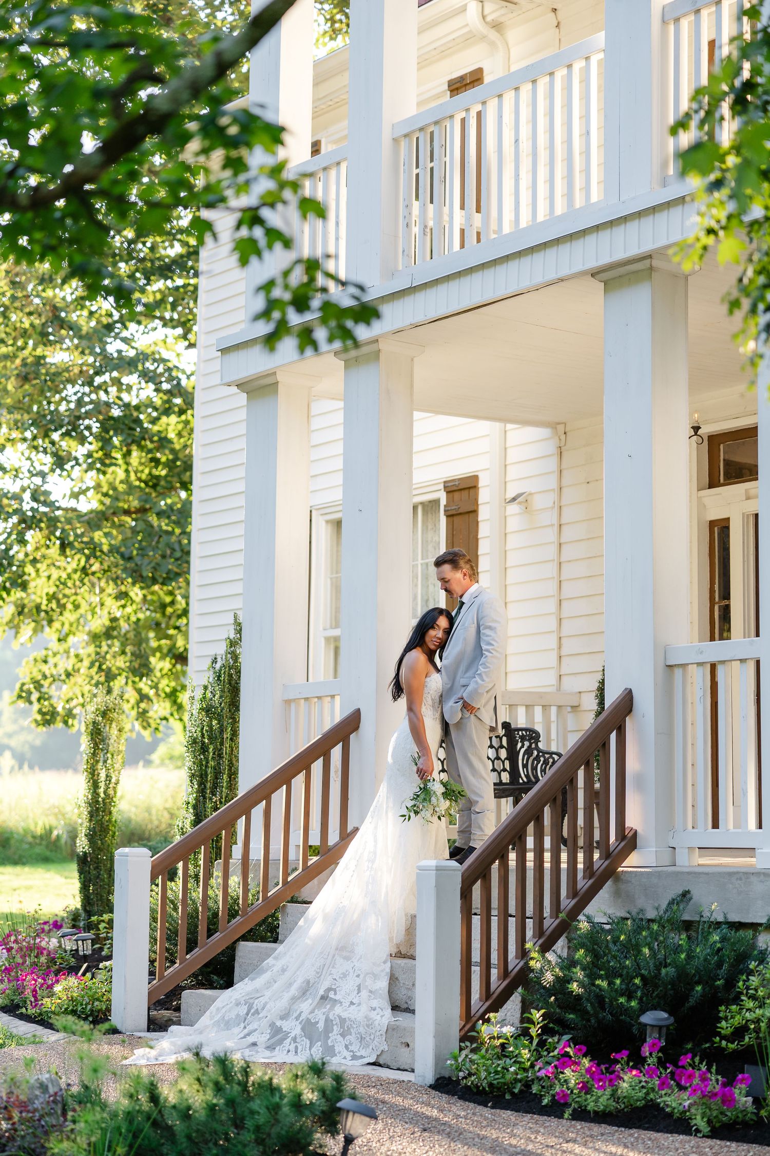 Bride and groom pose on white plantation house steps, bride in lace gown, surrounded by lush greenery.At Drakewood Farms in Nashville, TN