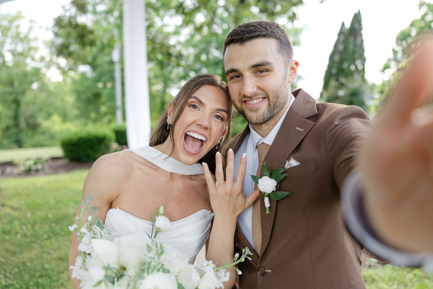 Happy bride and groom taking a wedding selfie, showing off engagement ring, bouquet, brown suit, white dress outdoors.