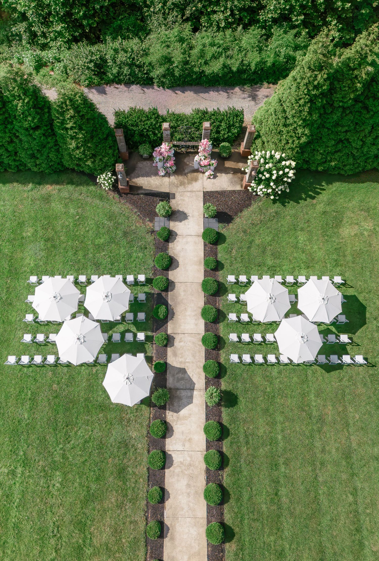 Aerial view of an elegant outdoor wedding ceremony setup with white chairs, umbrellas, and manicured hedges in a formal garden.