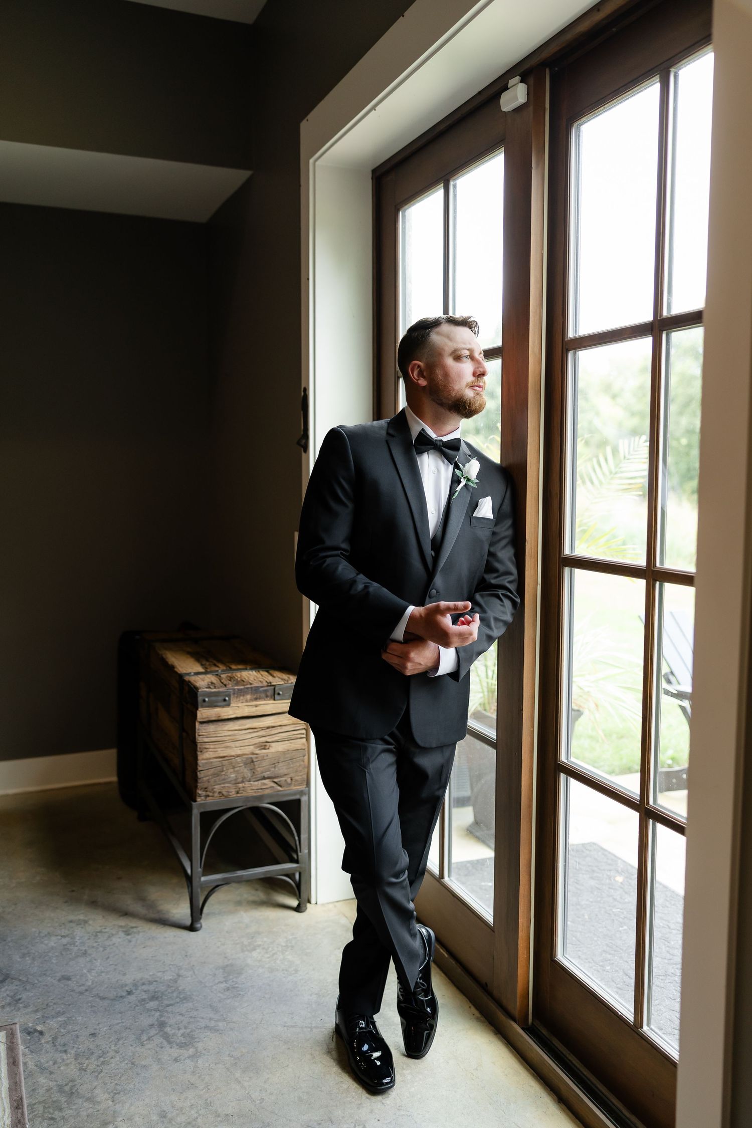 Groom in black tuxedo with bow tie stands by French doors, adjusting cufflinks before wedding ceremony. At The Venue at Birchwood