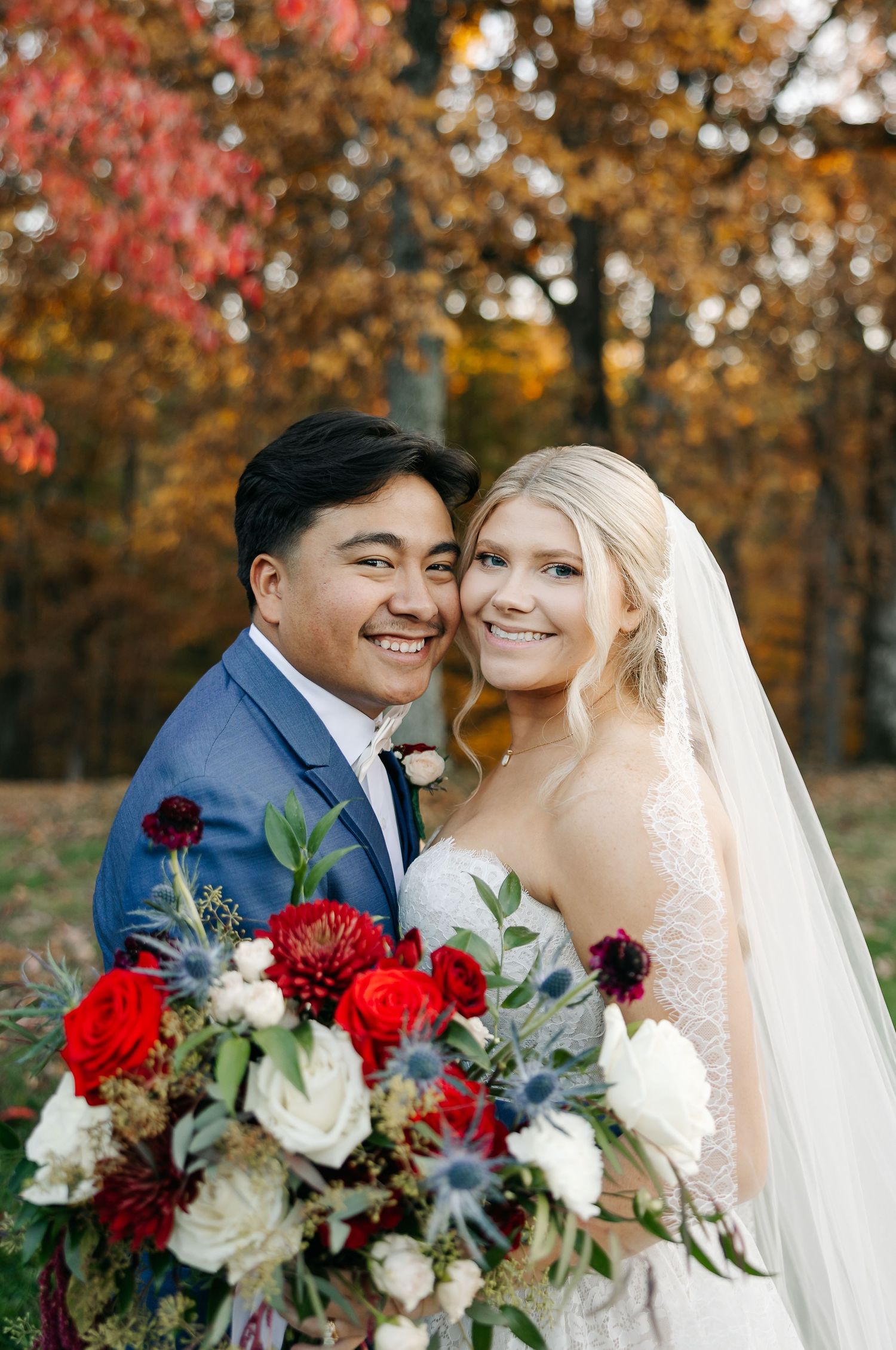 Smiling newlyweds pose outdoors in fall foliage, bride in lace gown with veil holding red and white bouquet, groom in blue suit.