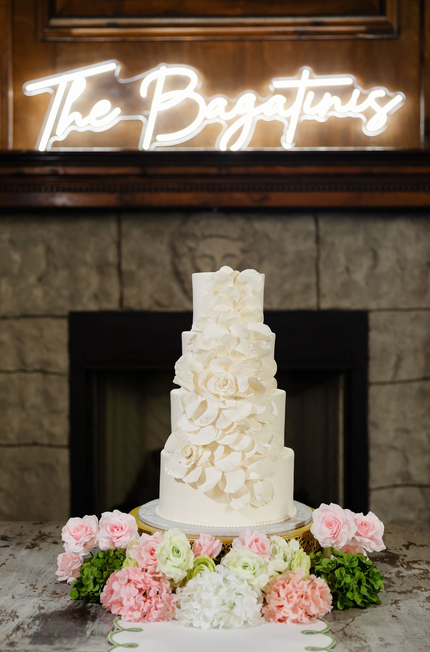 Elegant four-tier white wedding cake with ruffled petals, pink floral base, and neon sign reading The Bazatinis.