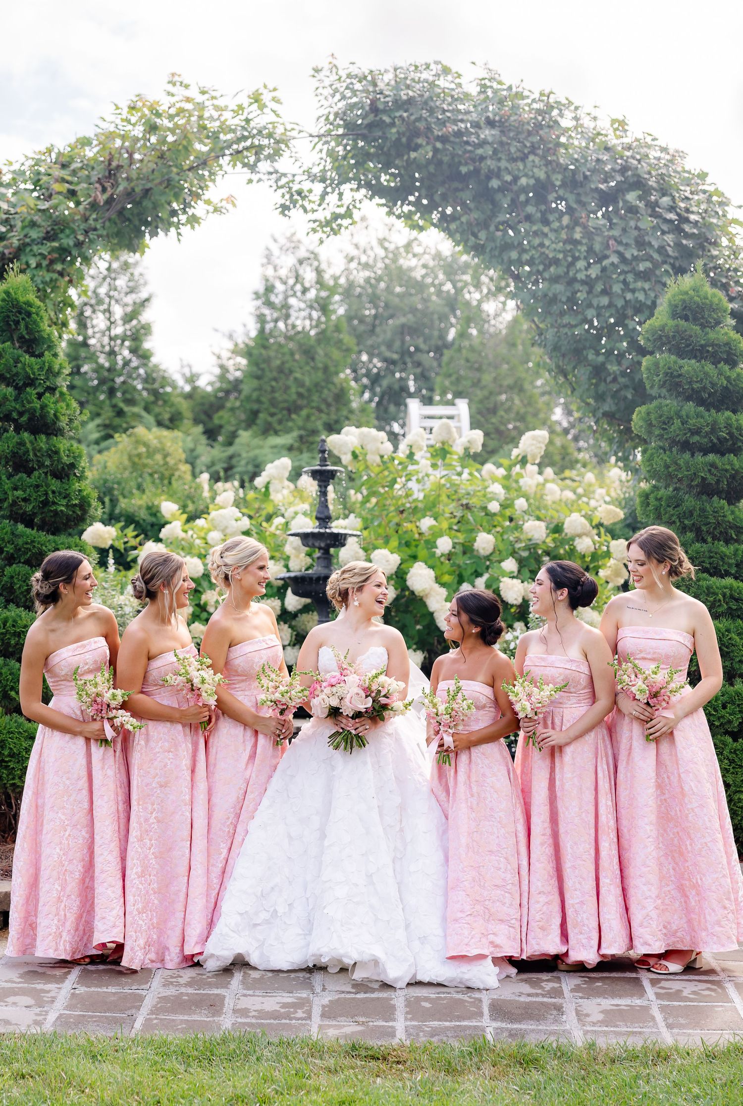 Bride in white gown with six bridesmaids in pink lace dresses holding bouquets in a lush garden with floral arch.