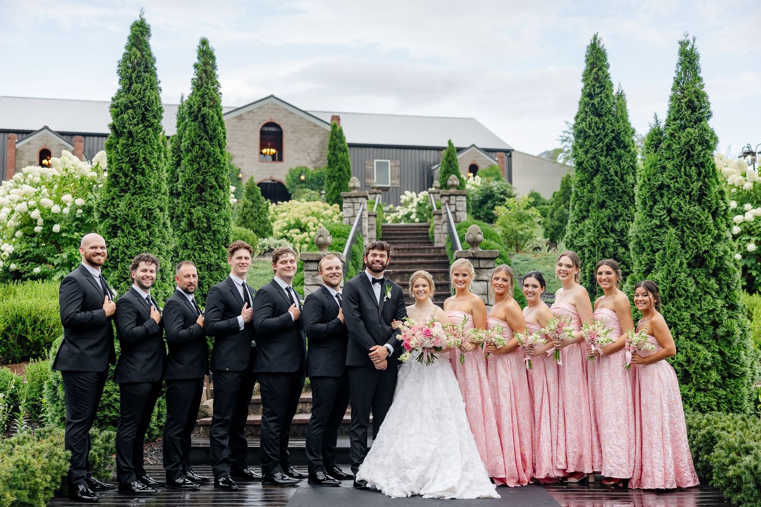 Wedding party poses outdoors with bride in lace gown, groomsmen in black tuxedos, and bridesmaids in pink dresses near tall hedges.