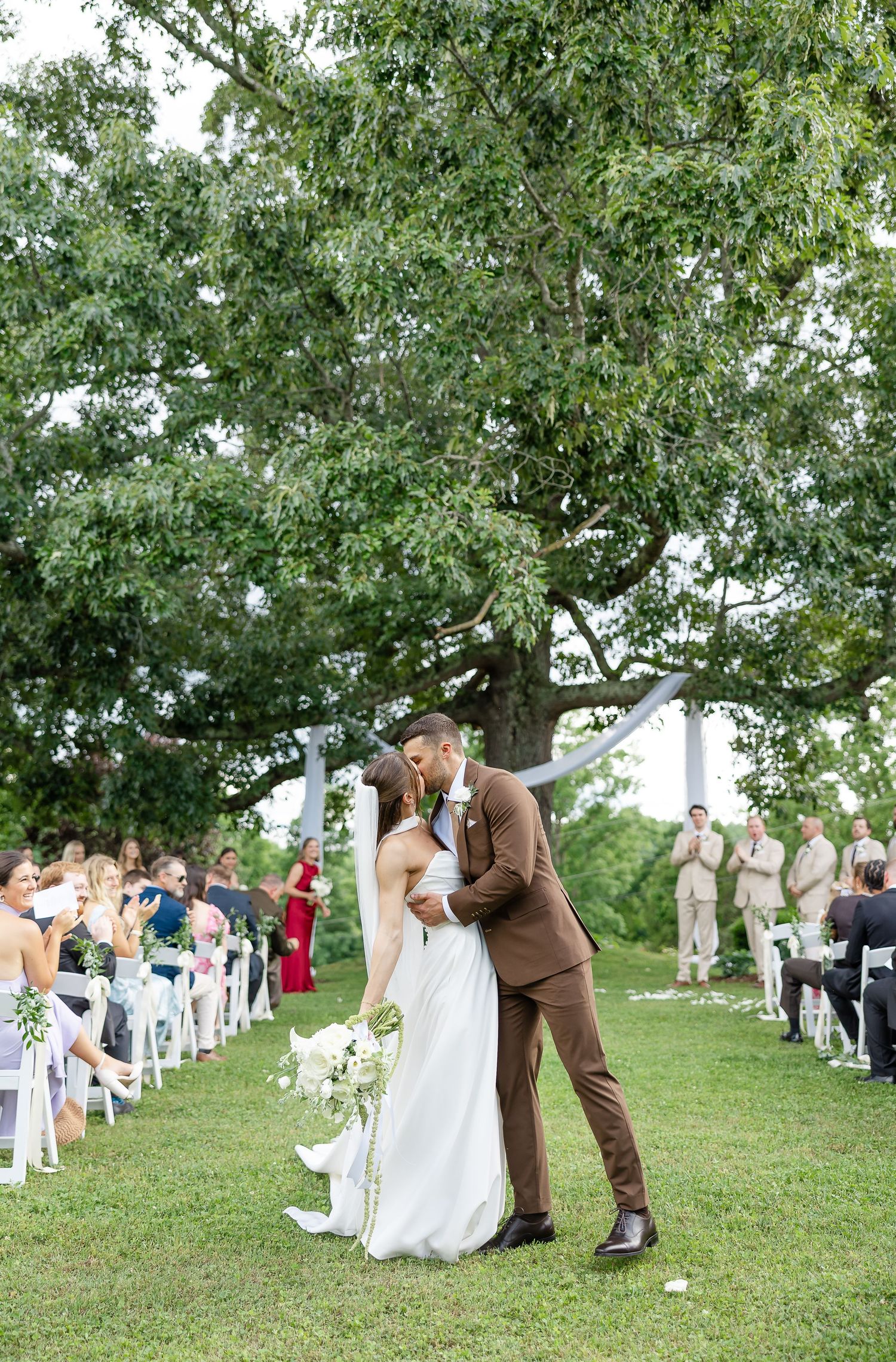 Bride and groom share first kiss at outdoor wedding ceremony under large tree with white chairs lining the aisle.