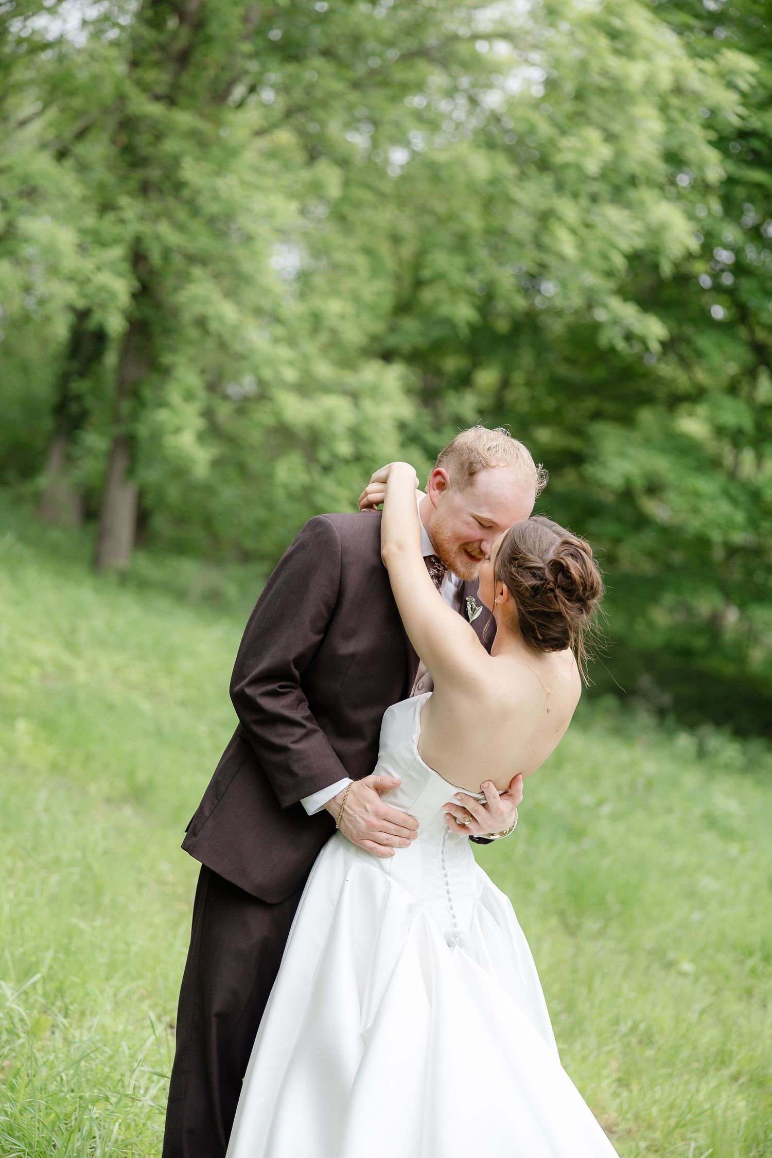 A groom in a dark suit kisses his bride in a backless white gown outdoors in a lush green garden.