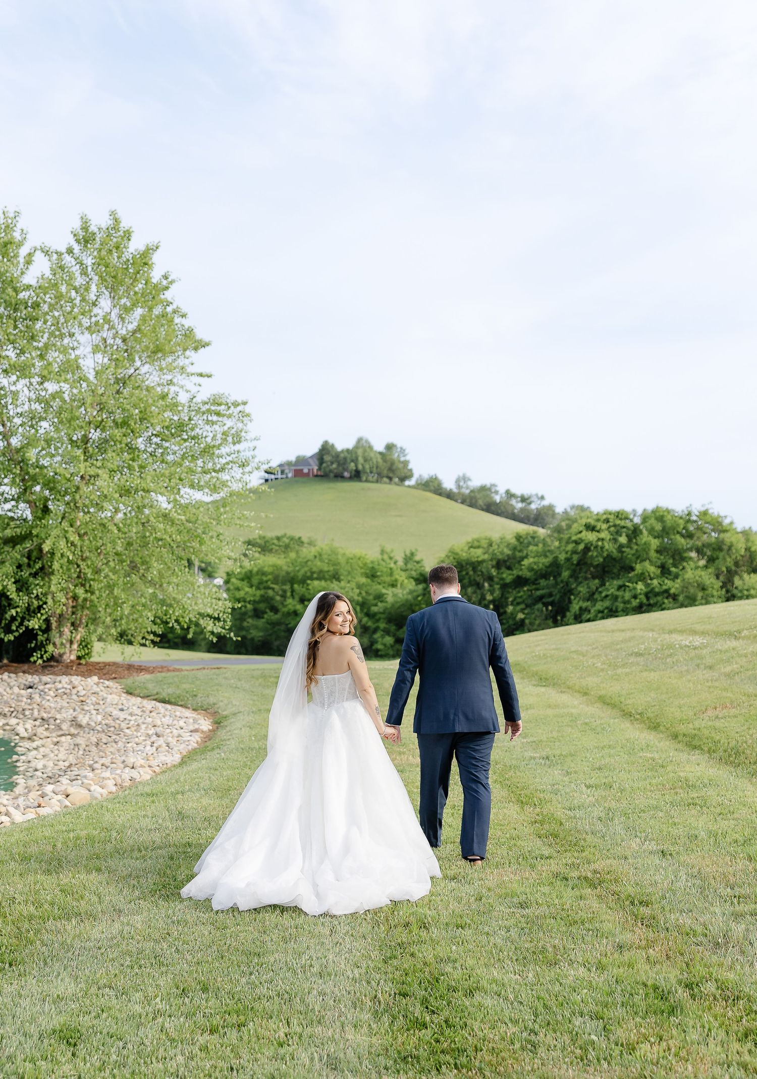 Bride and groom walking hand in hand across lush green rolling hills on their wedding day.