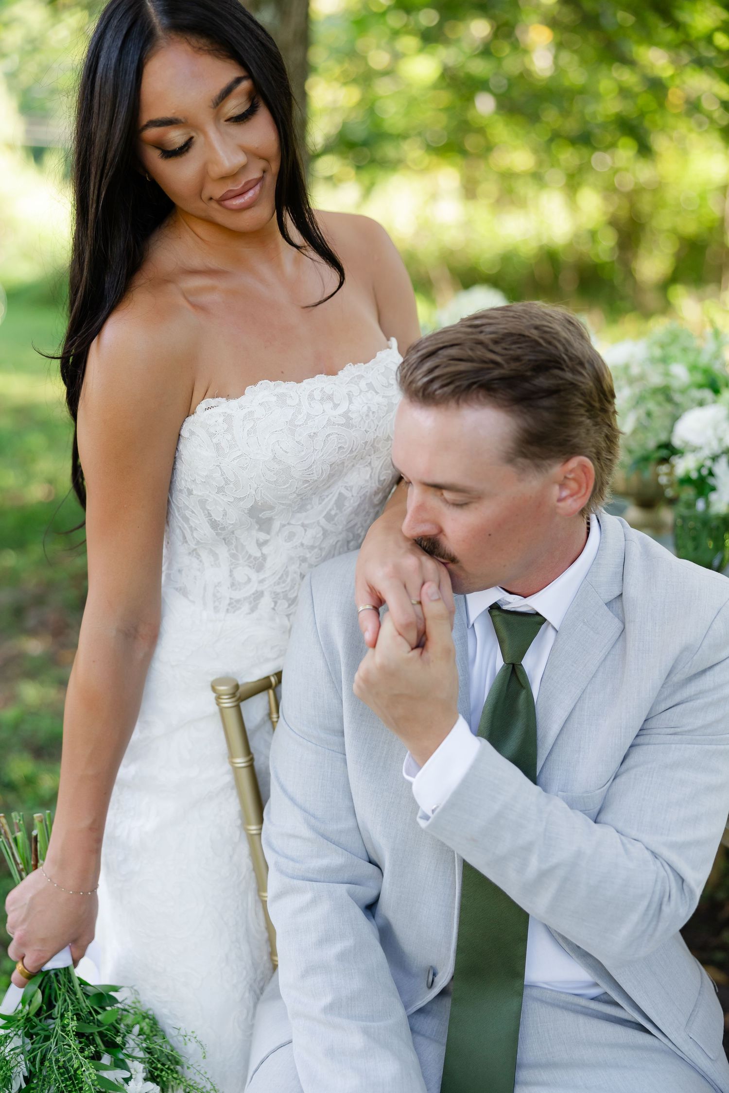 Groom in light gray suit kisses bride's hand as she smiles in white lace wedding dress outdoors. At Drakewood Farms in Nashville, TN