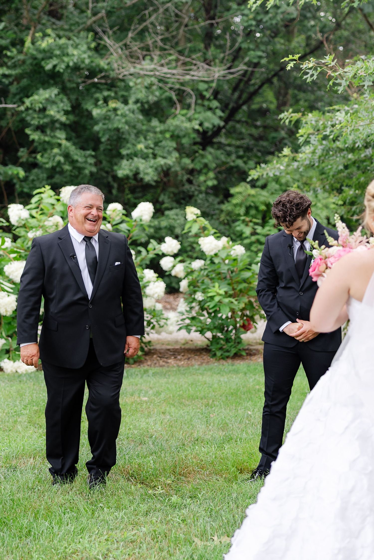 Joyful father of the bride smiling during outdoor wedding ceremony with white hydrangeas and lush greenery backdrop.