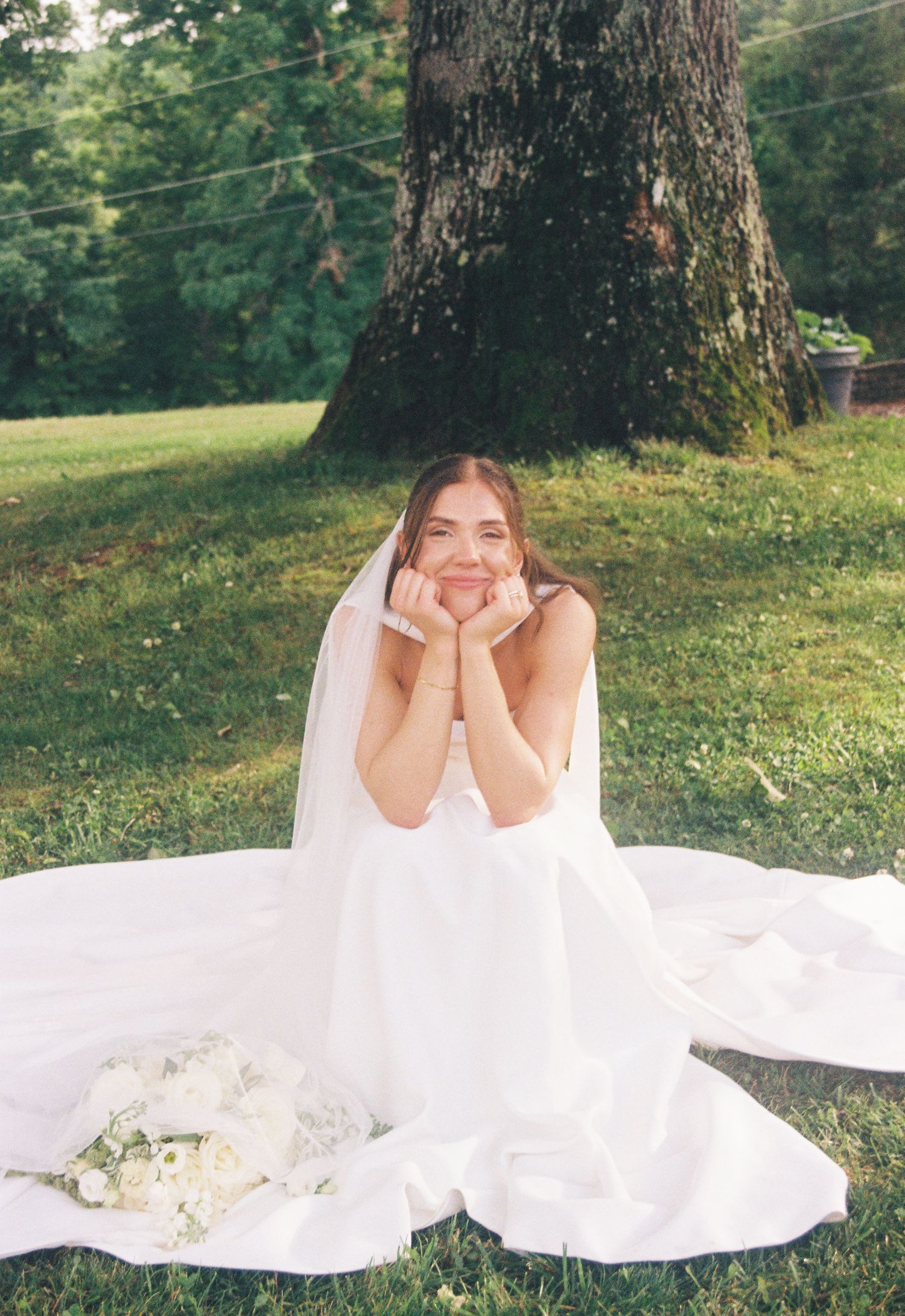 Smiling bride in white gown and veil sitting on grass near tree with bouquet of white flowers.