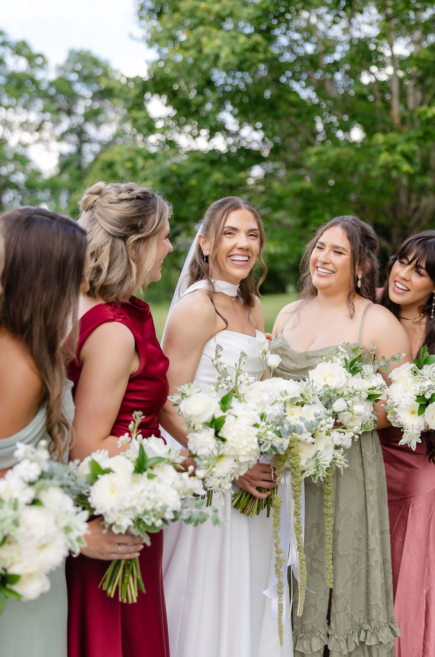 Bride in white gown laughs with bridesmaids in colorful dresses holding white floral bouquets outdoors.