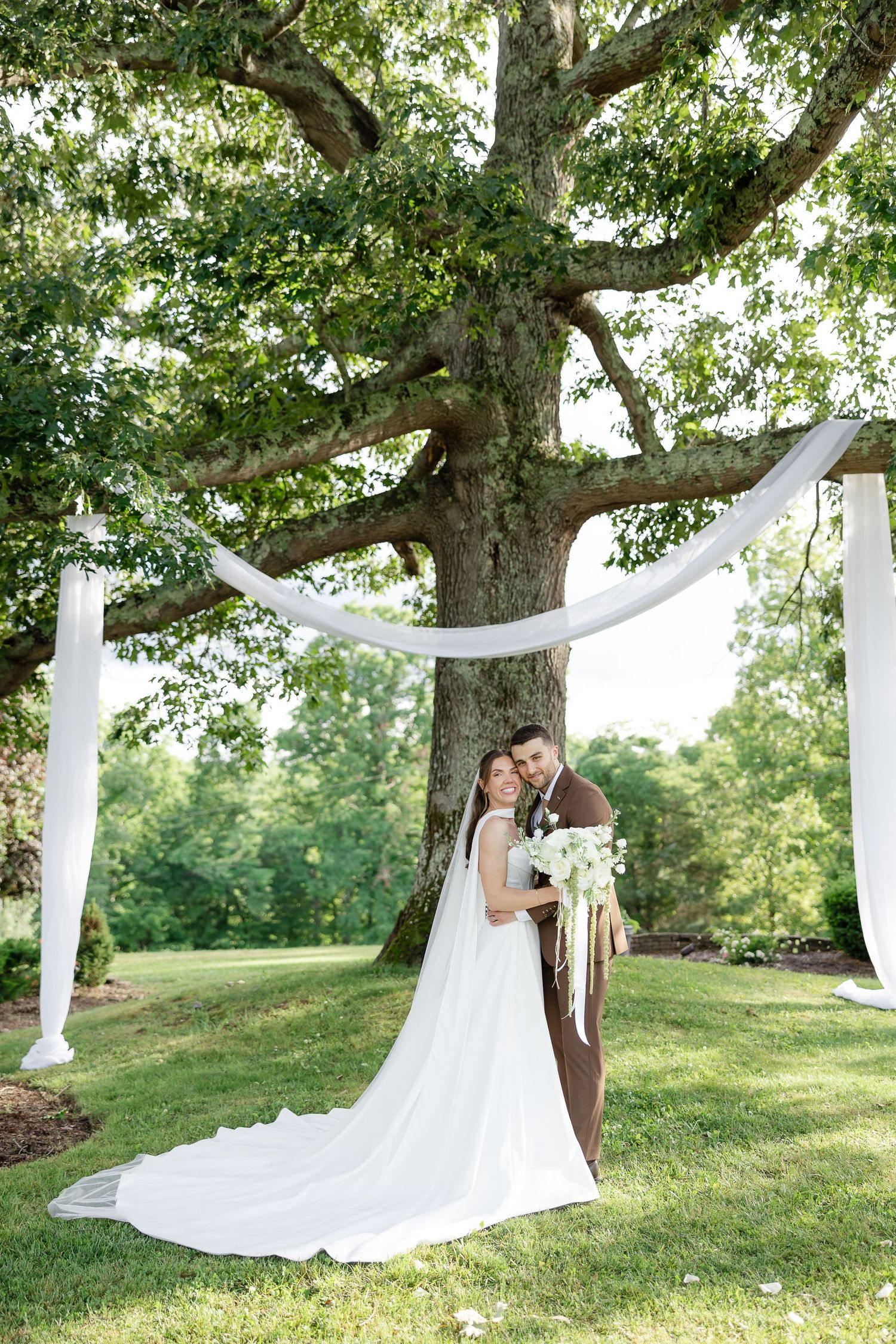 Bride and groom pose under a grand oak tree with white draping at an outdoor wedding ceremony.