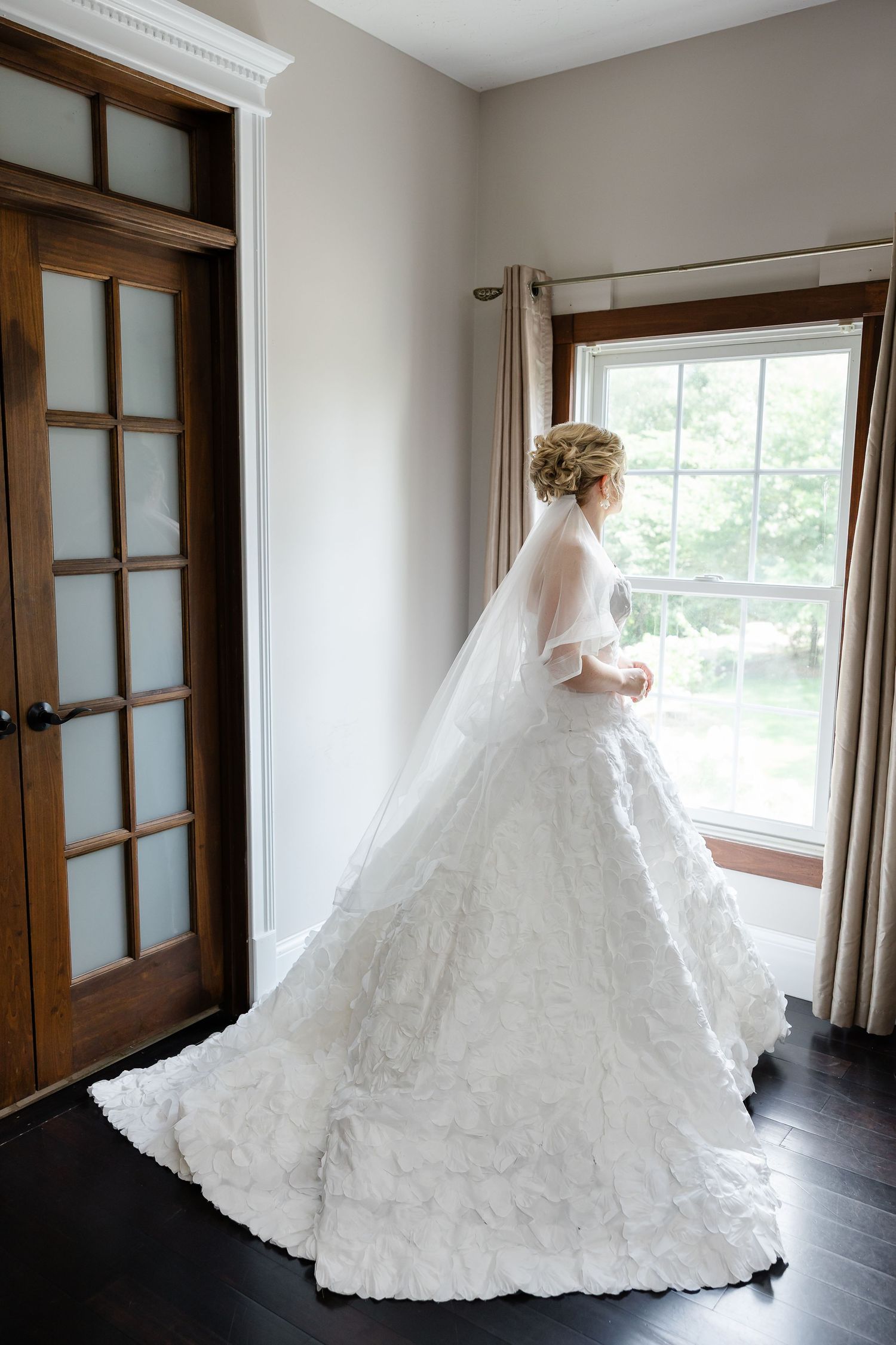 Bride in elegant white lace ball gown with veil stands by window in softly lit room with dark hardwood floors.