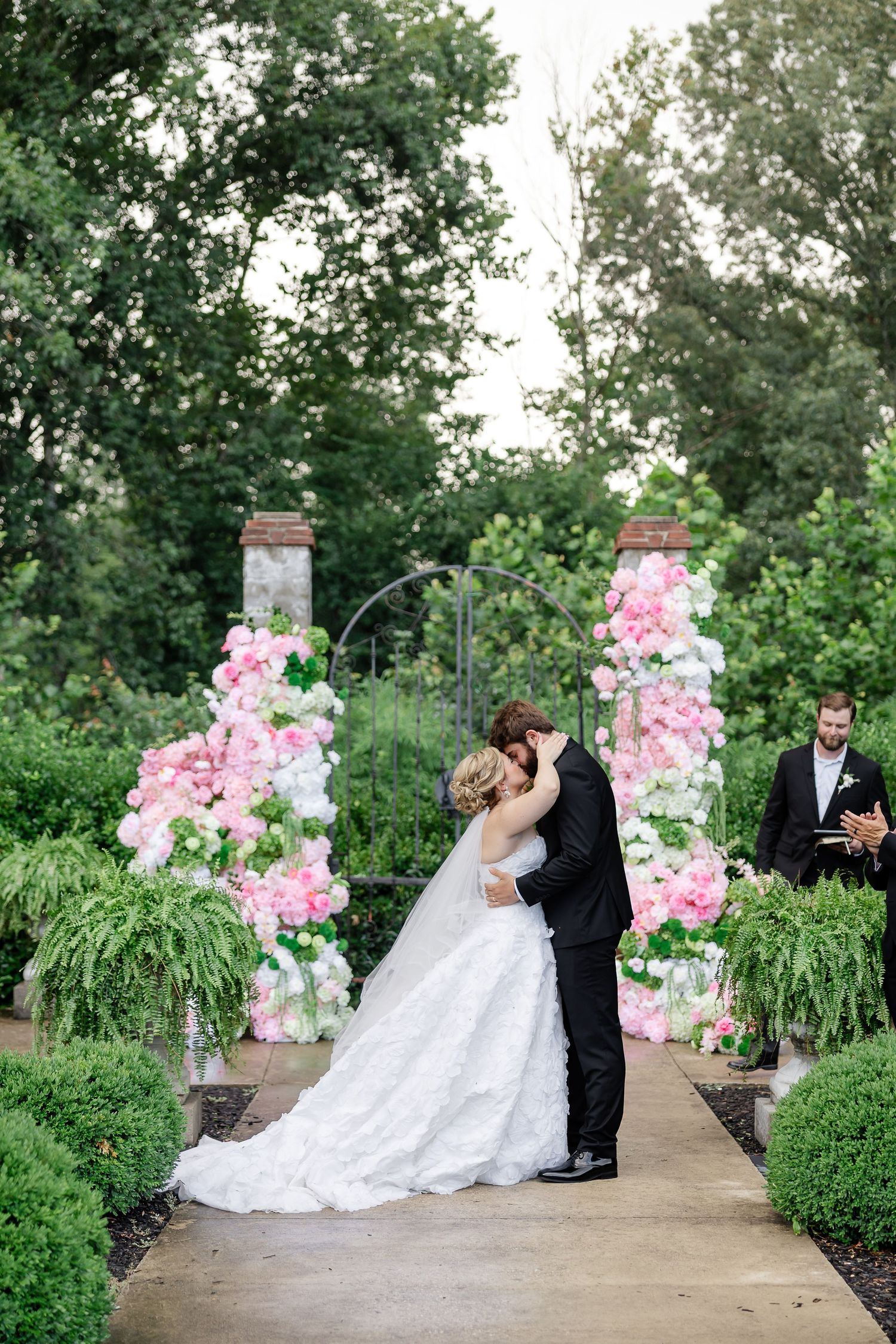 Bride and groom share first kiss at outdoor garden wedding ceremony with pink and white floral columns.