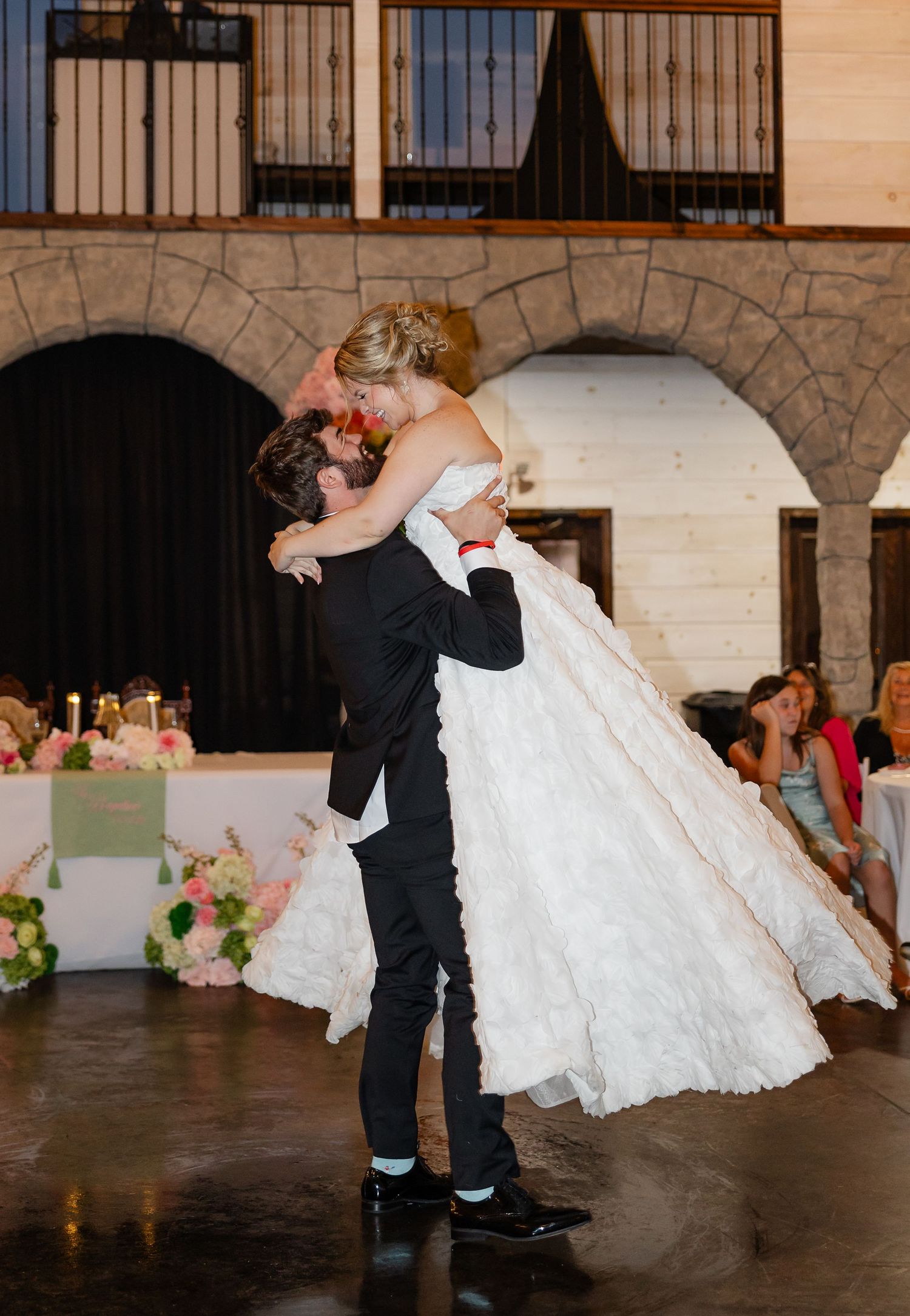 Groom lifts bride in white wedding gown during first dance at elegant stone venue reception.