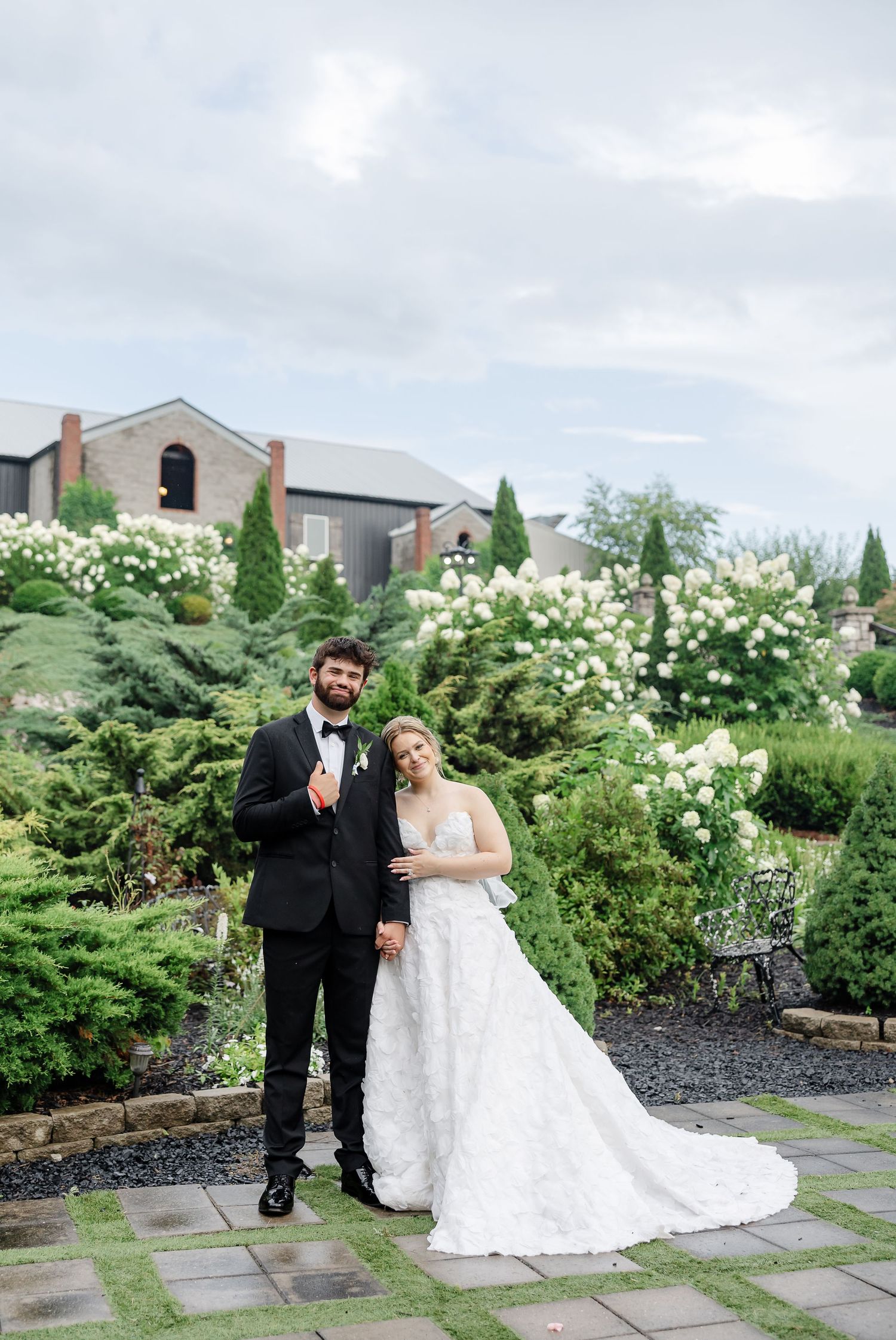 Bride and groom pose in lush garden with white florals and stone building backdrop on their wedding day.