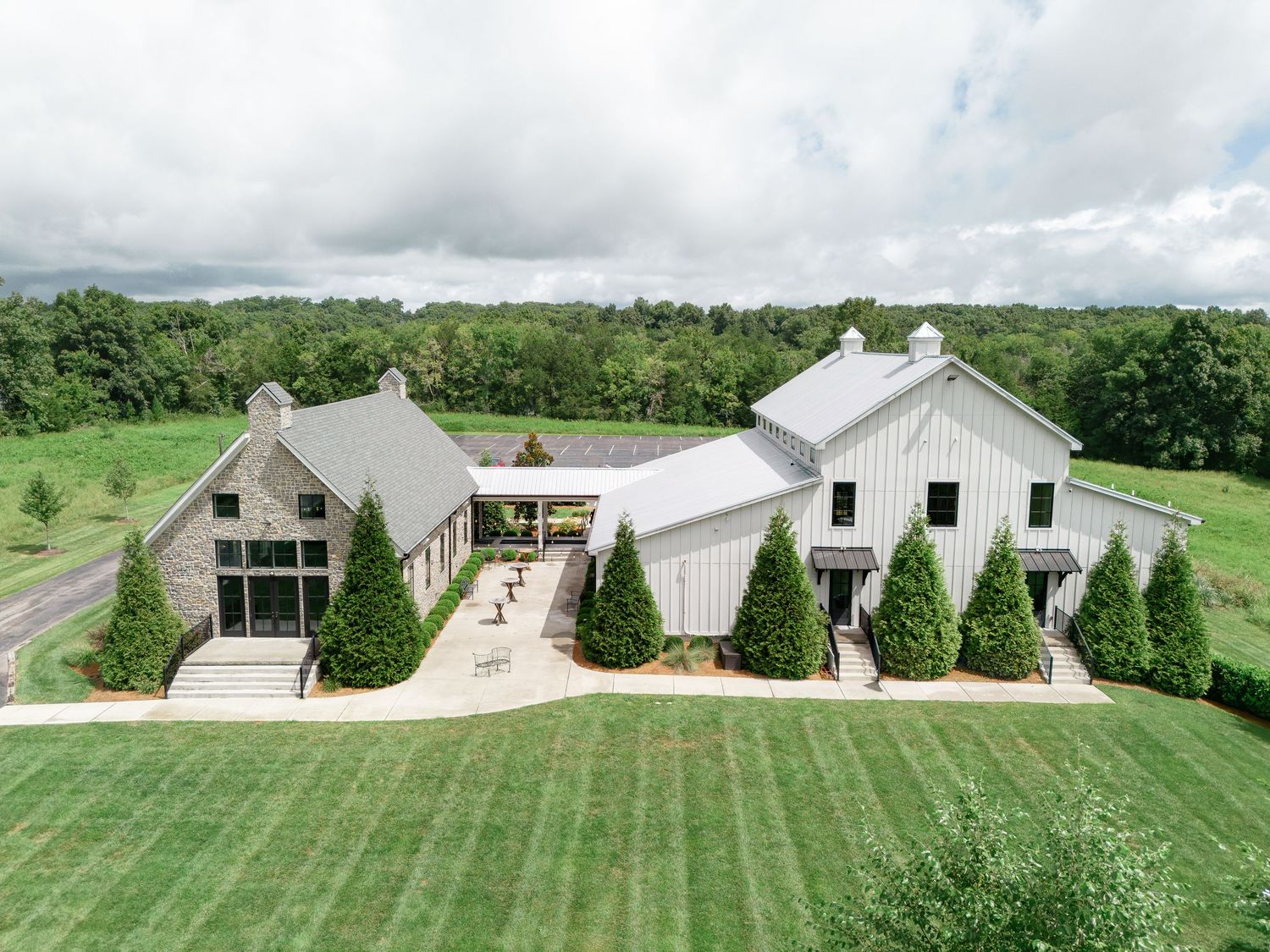 Aerial view of a modern white barn wedding venue surrounded by lush green fields and trees. At The Venue at Birchwood