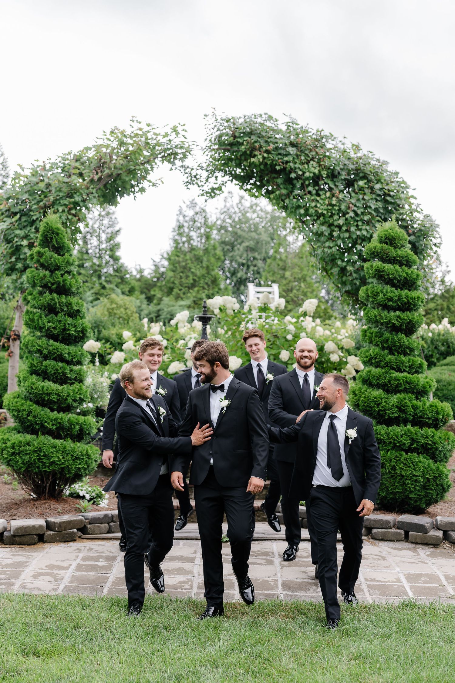 Groom and groomsmen in black tuxedos walking through a lush garden archway at an elegant outdoor wedding.