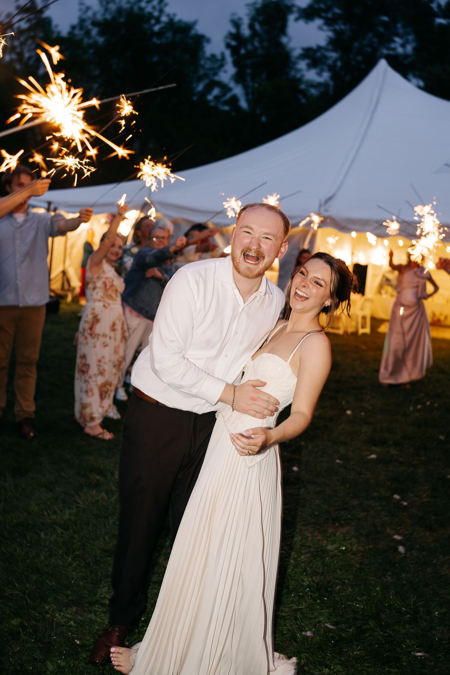 Joyful newlyweds embrace during a sparkler send-off at an outdoor evening wedding reception under a white tent.