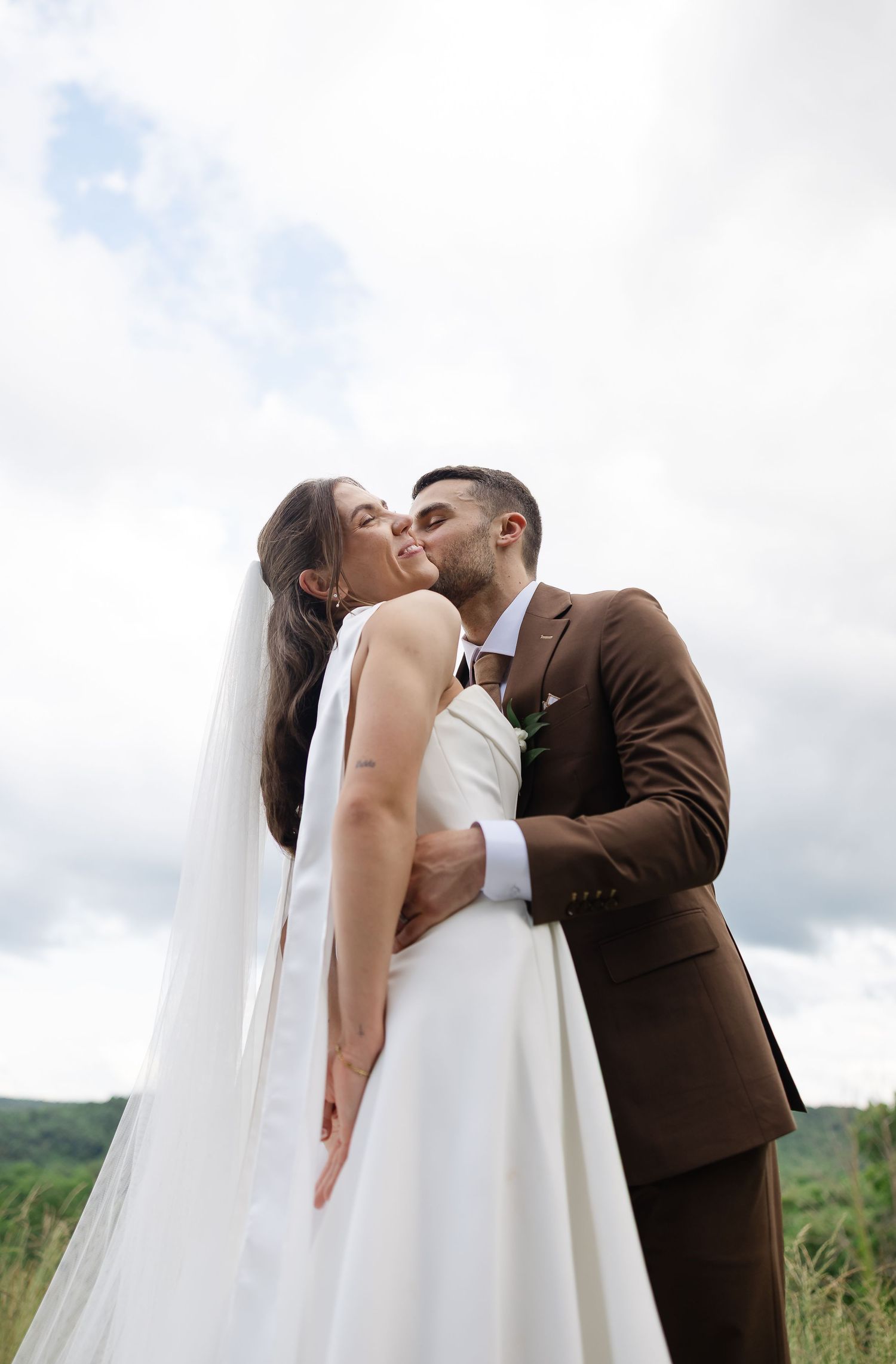 A bride and groom share a romantic kiss outdoors, with the bride in a white gown and veil and groom in a brown suit.