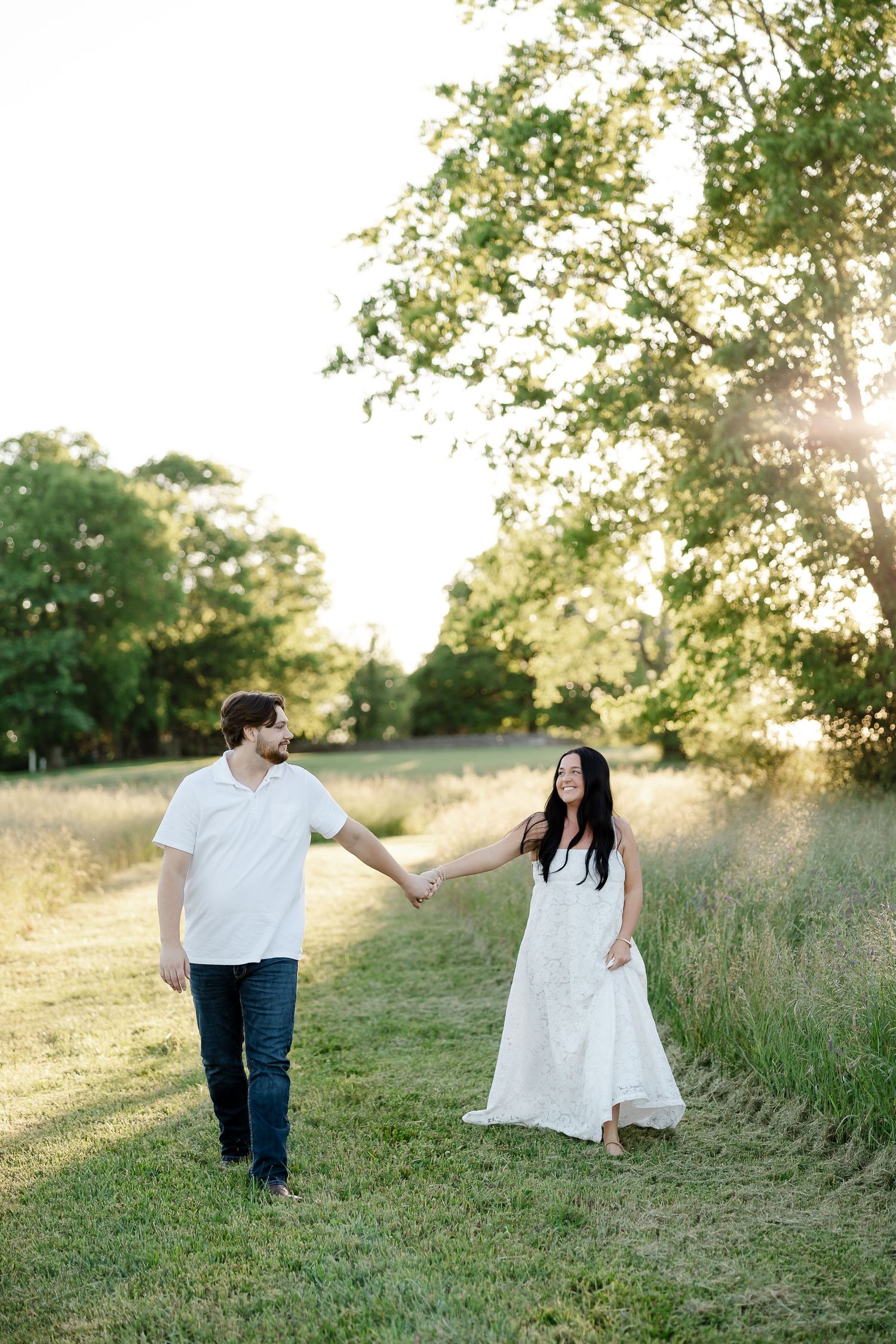 Couple holding hands walking through a sunny green field at golden hour, woman in white dress, man in white shirt and jeans.
