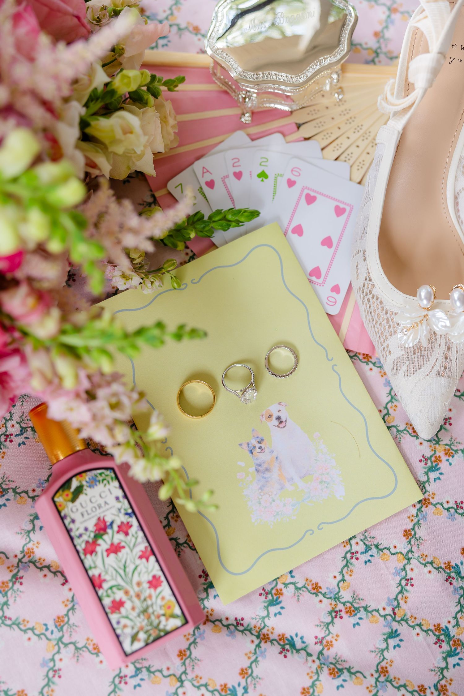 Wedding flat lay with gold rings on yellow journal, playing cards, pink fan, floral bouquet, Gucci perfume, and bridal heels.